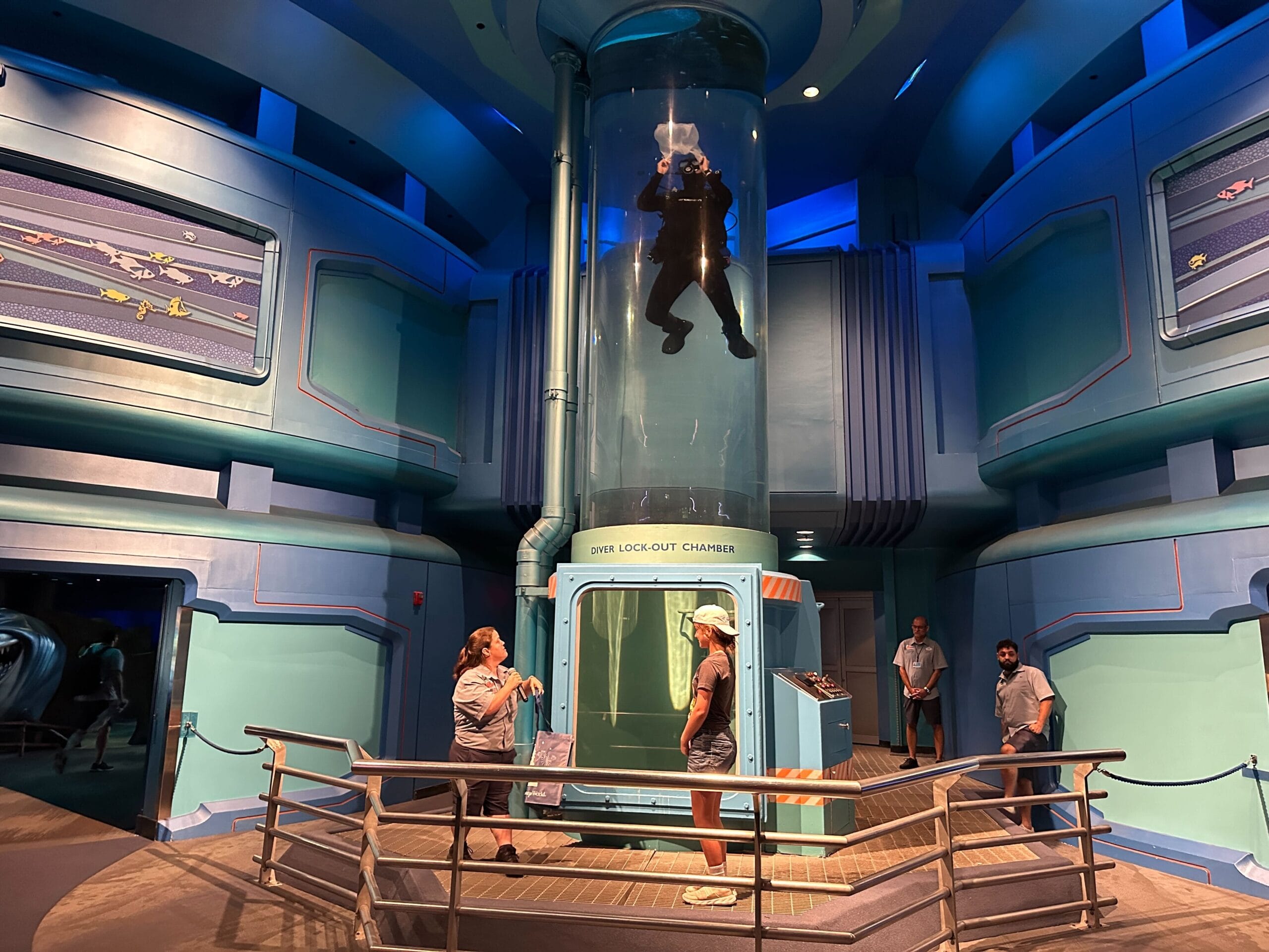 A person in a wetsuit floats in a diver lock-out chamber demo as others watch in a blue, futuristic-themed room.