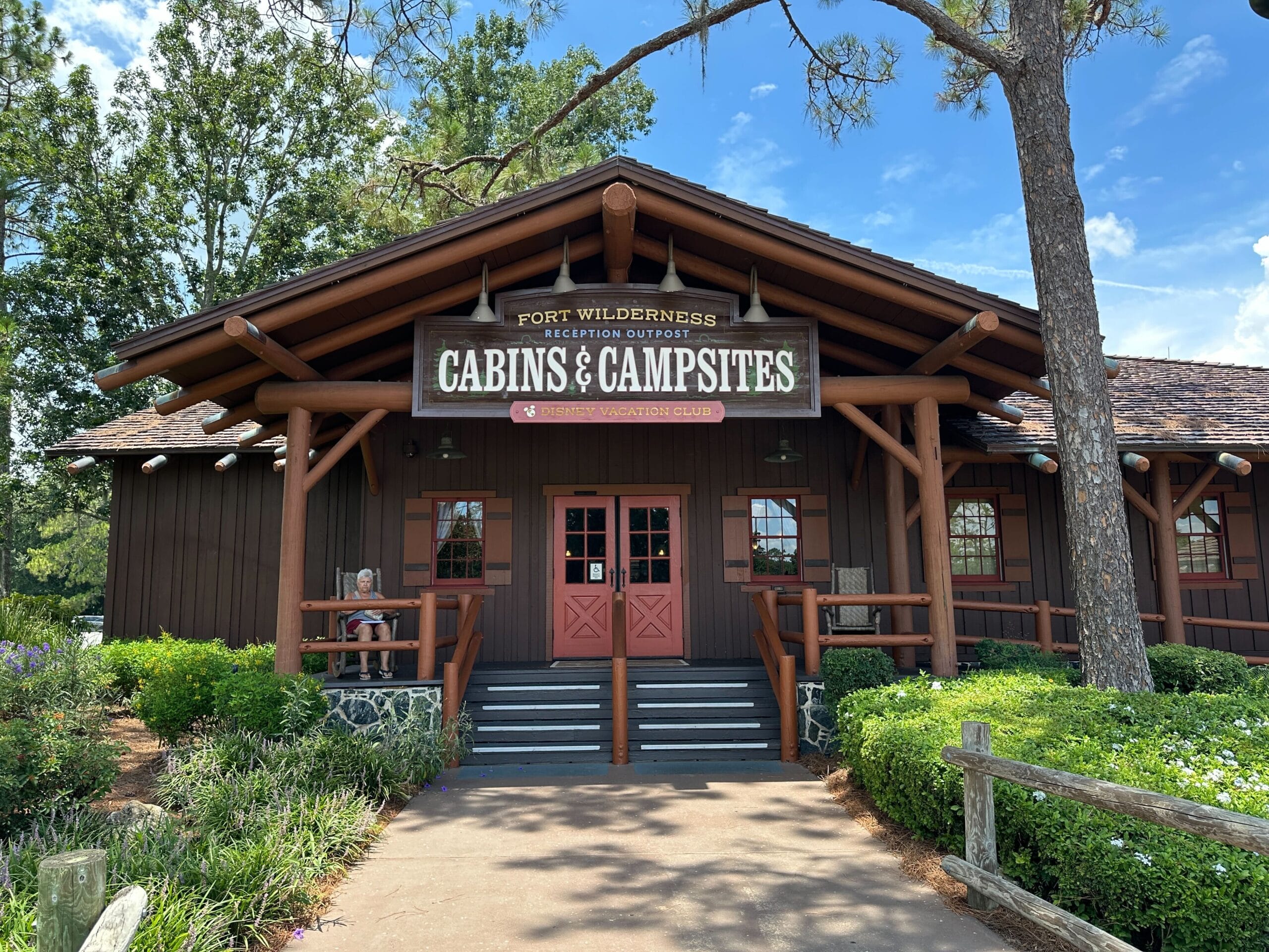 Rustic wooden building with a sign reading “Cabins & Campsites” at Fort Wilderness under a blue sky with greenery.