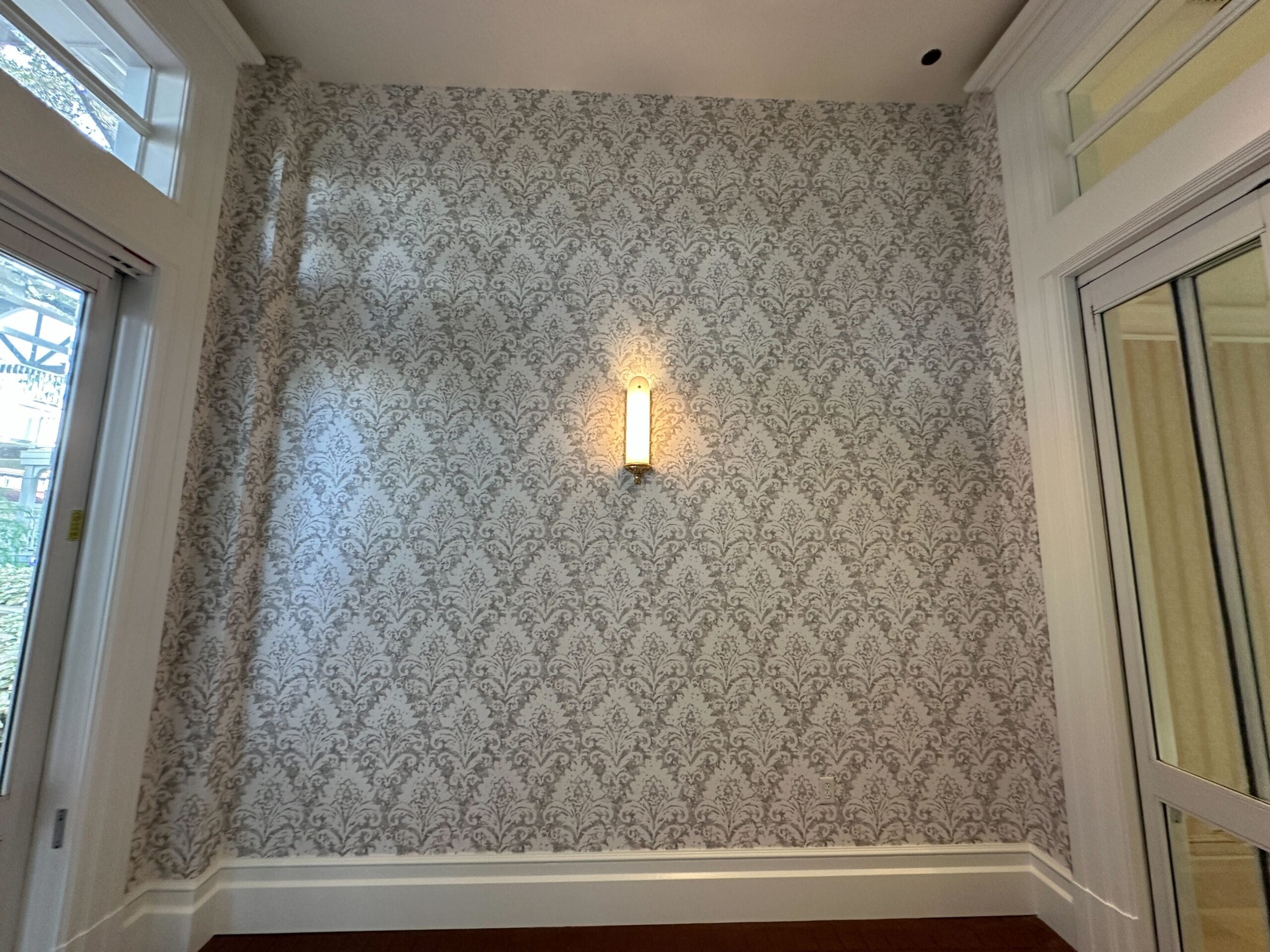 A wall with ornate patterned wallpaper and a single sconce light, next to windows and a glass door.