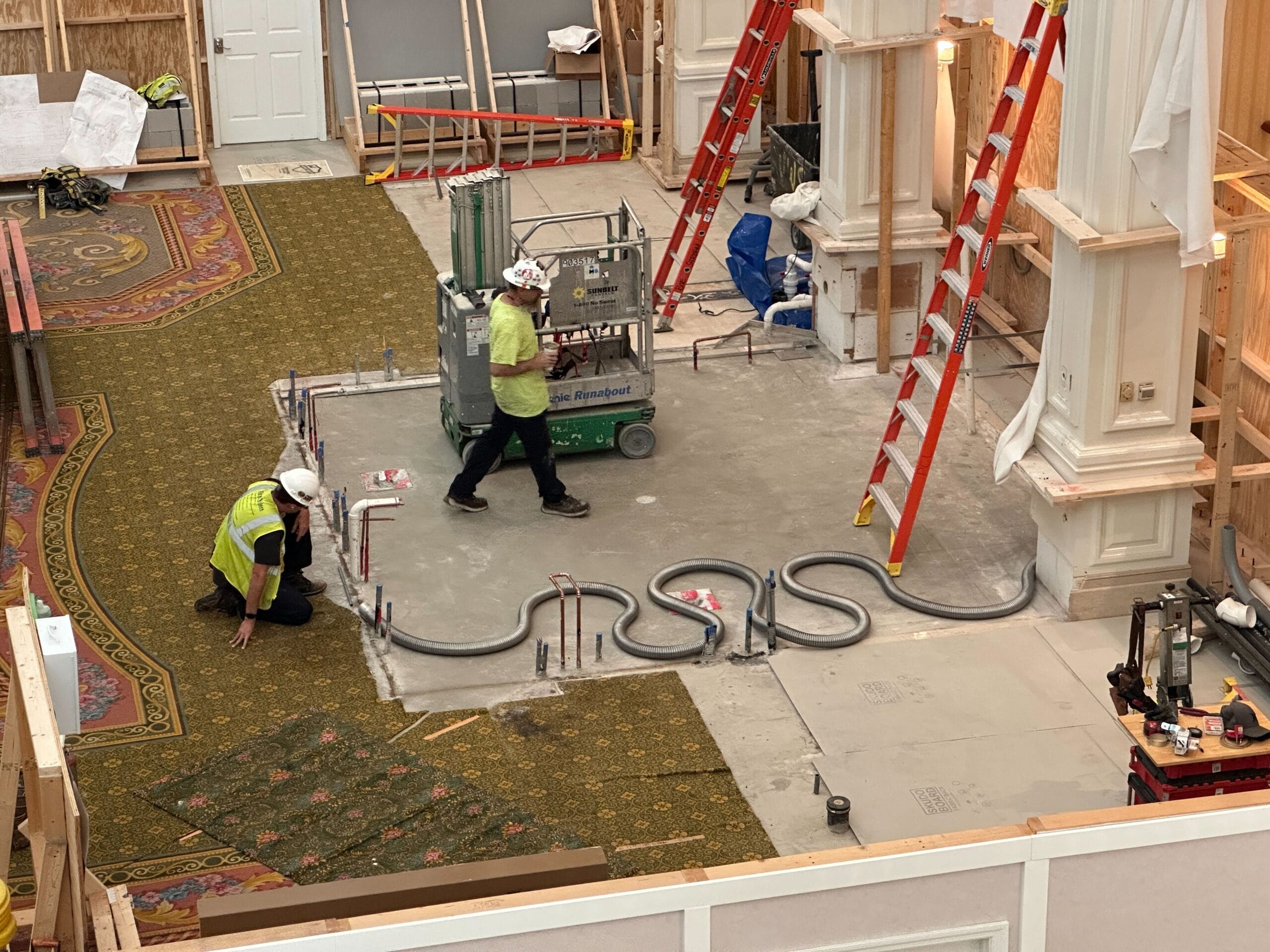 Two construction workers install electrical conduit on the floor of a partially finished building with carpet and tools around.