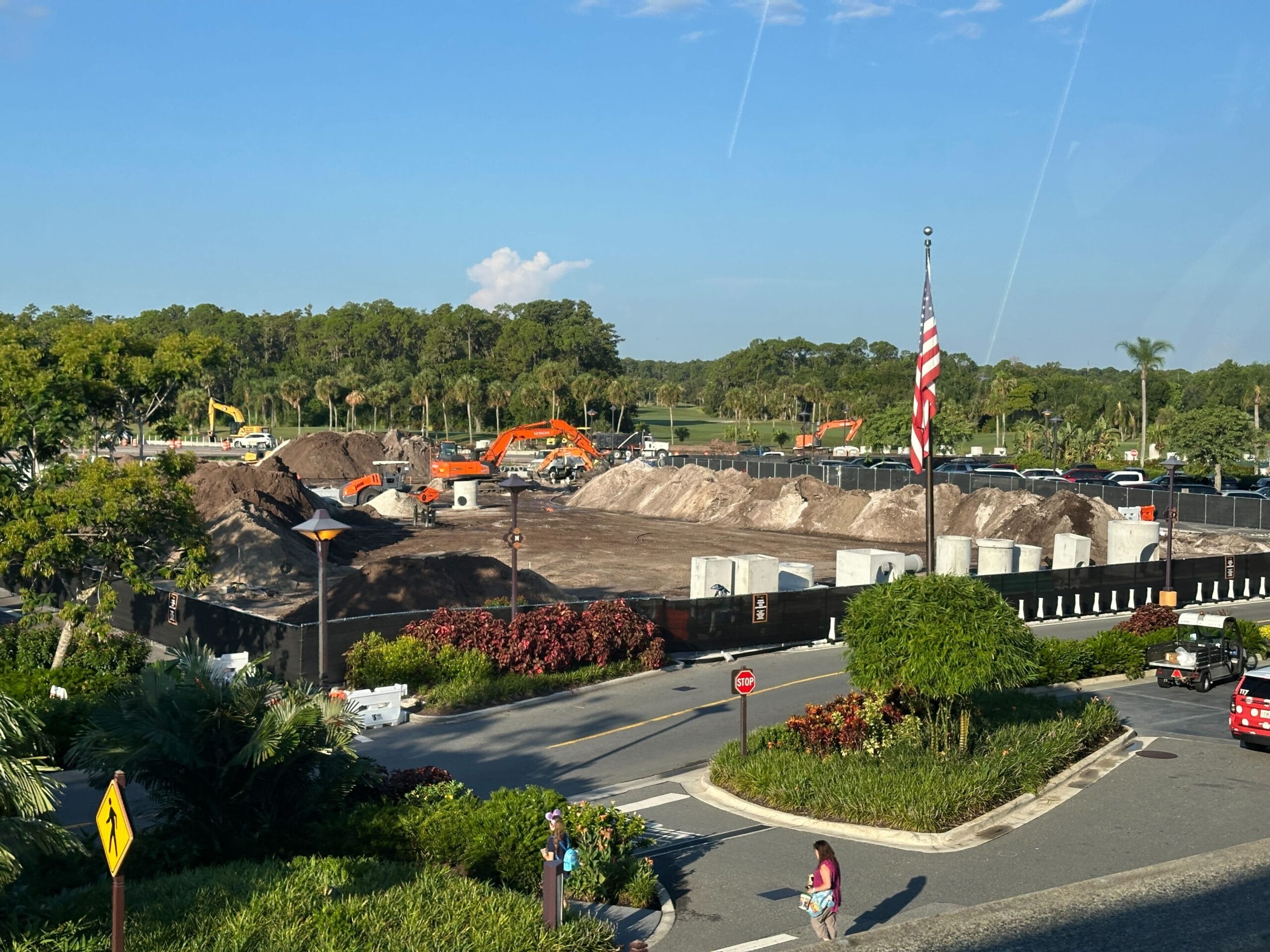 A construction site with excavators, dirt piles, and an American flag beside a road with cars and pedestrians.