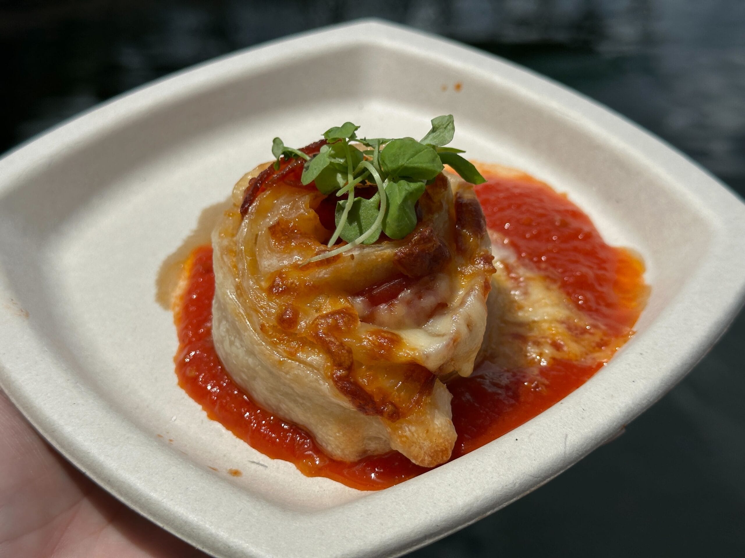 A New Food & Wine savory pastry roll topped with melted cheese and greens, served on tomato sauce in a square dish.