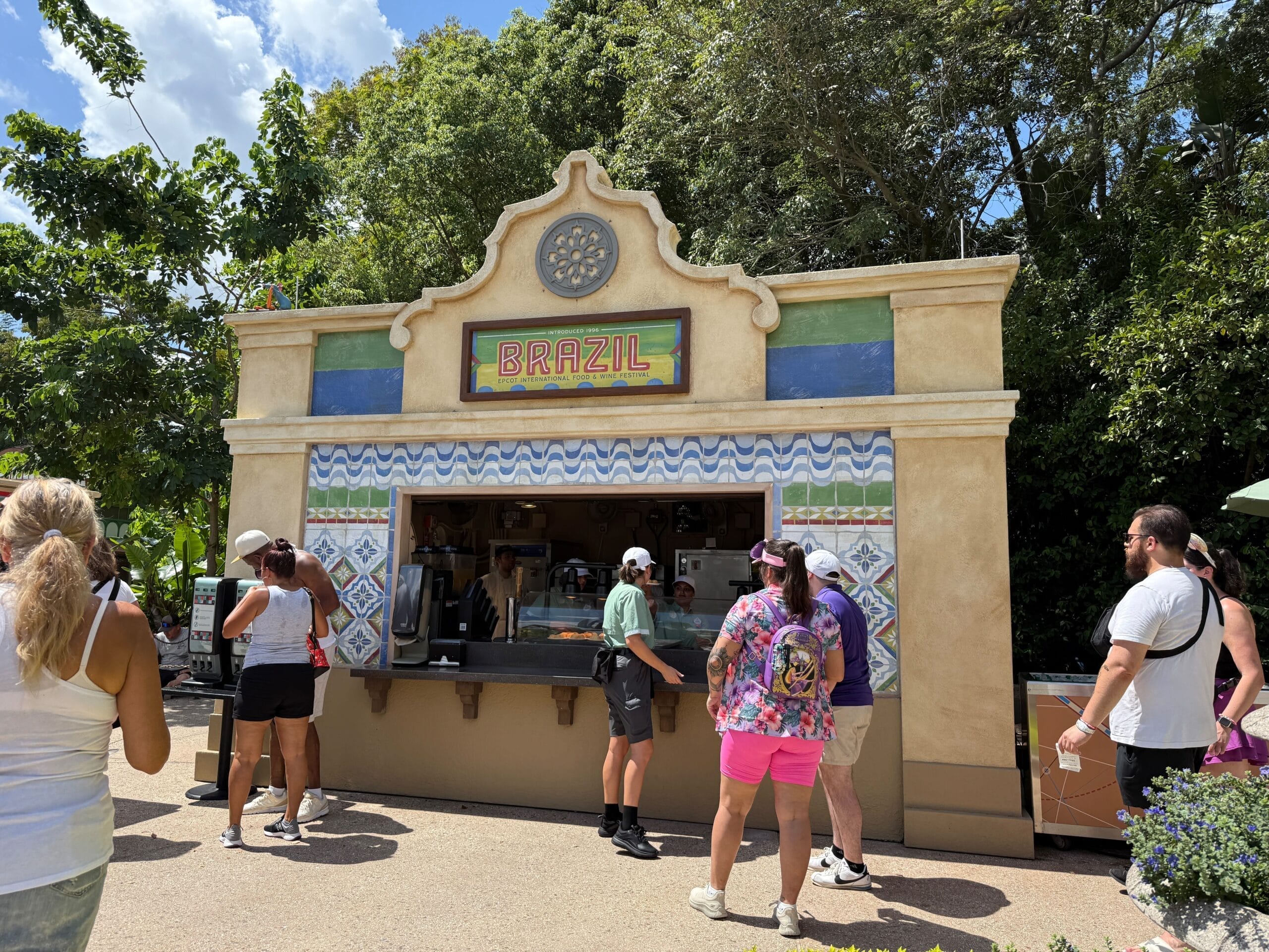 People stand in line at a Brazil-themed EPCOT food booth during the 2025 Food and Wine Festival, surrounded by trees.