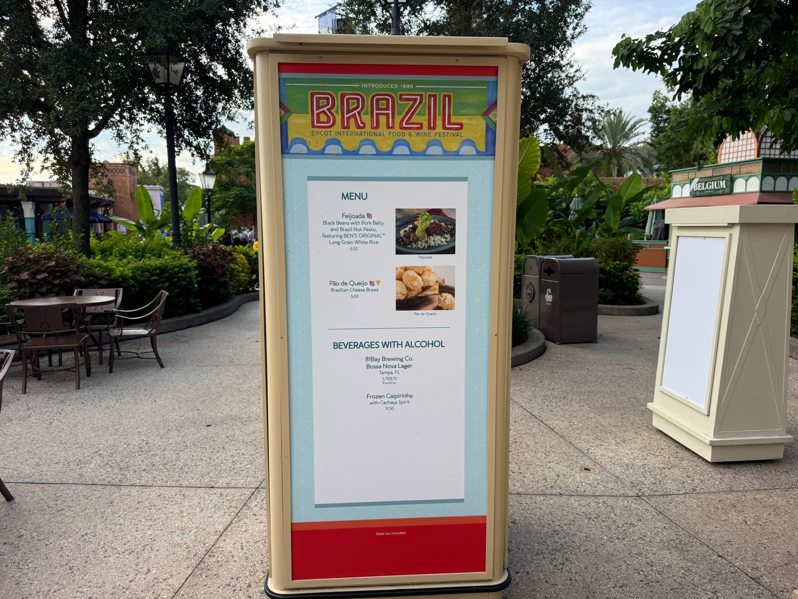 Outdoor Brazil Food & Wine menu sign listing dishes and drinks at a festival, surrounded by greenery and patio seating.