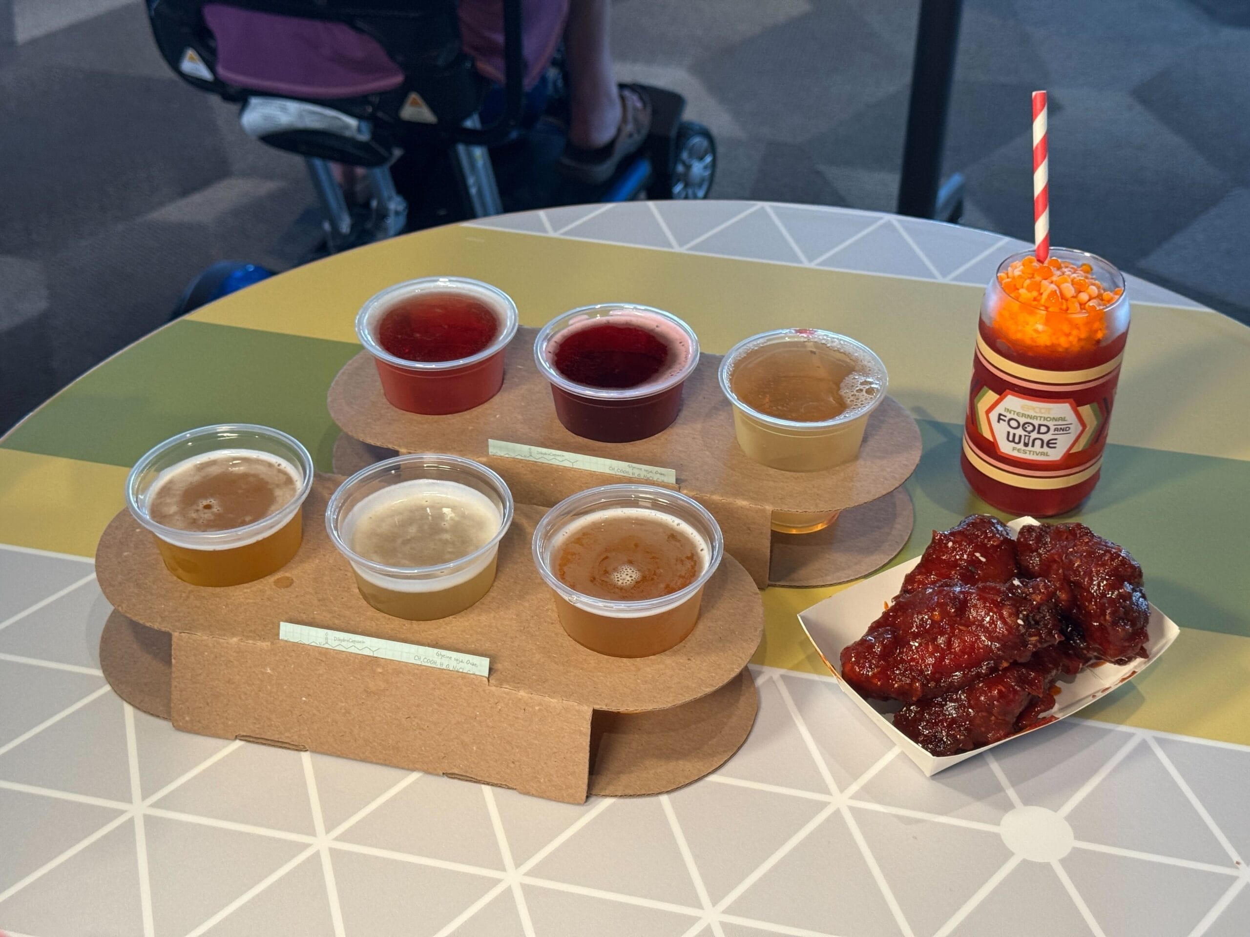 A beer flight, sticky glazed chicken wings, and a specialty drink at the EPCOT Food and Wine Festival on a vibrant table.