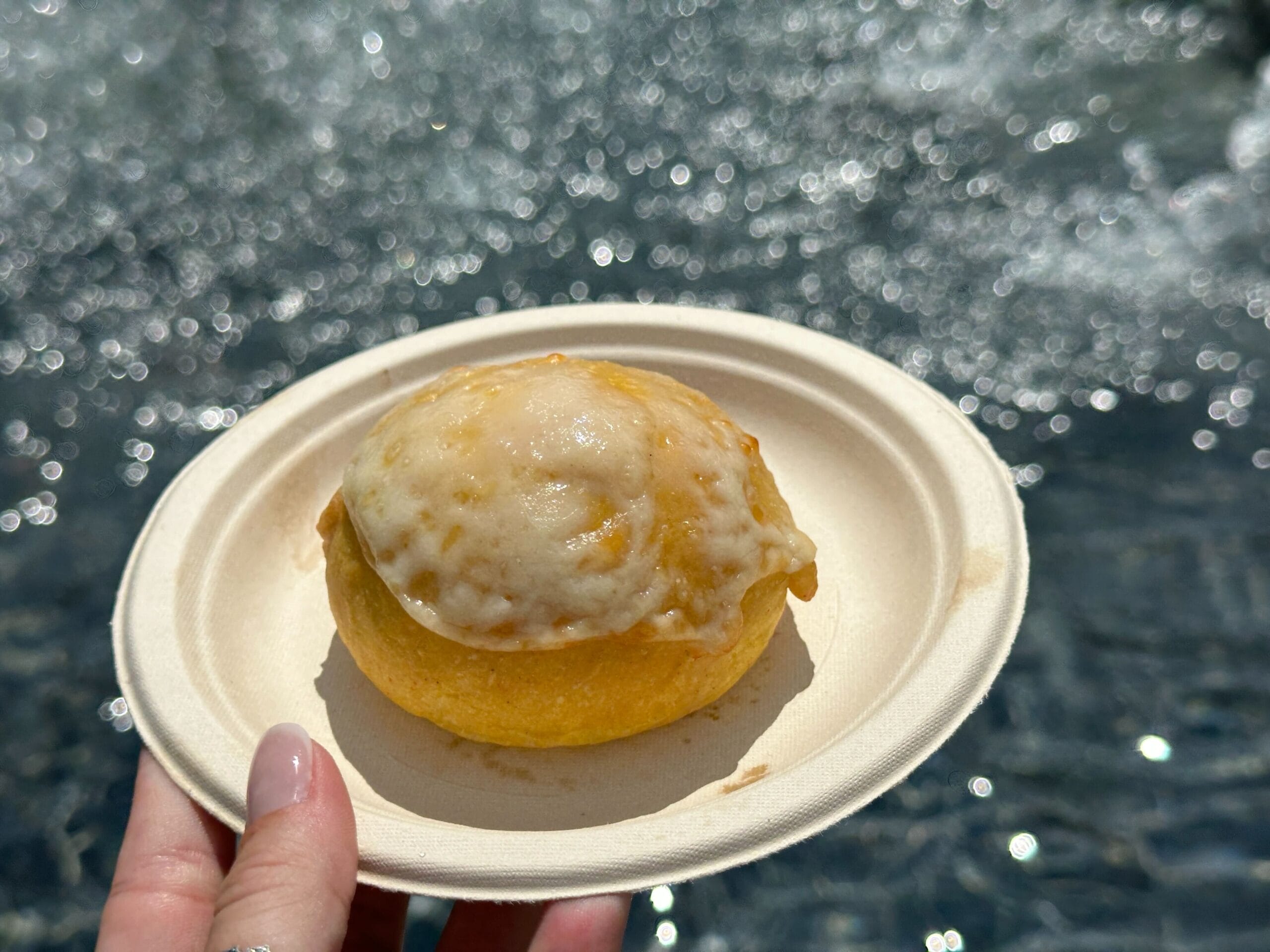 A hand holds a cheese-topped pastry at the EPCOT Food and Wine Festival against a sparkling water backdrop.