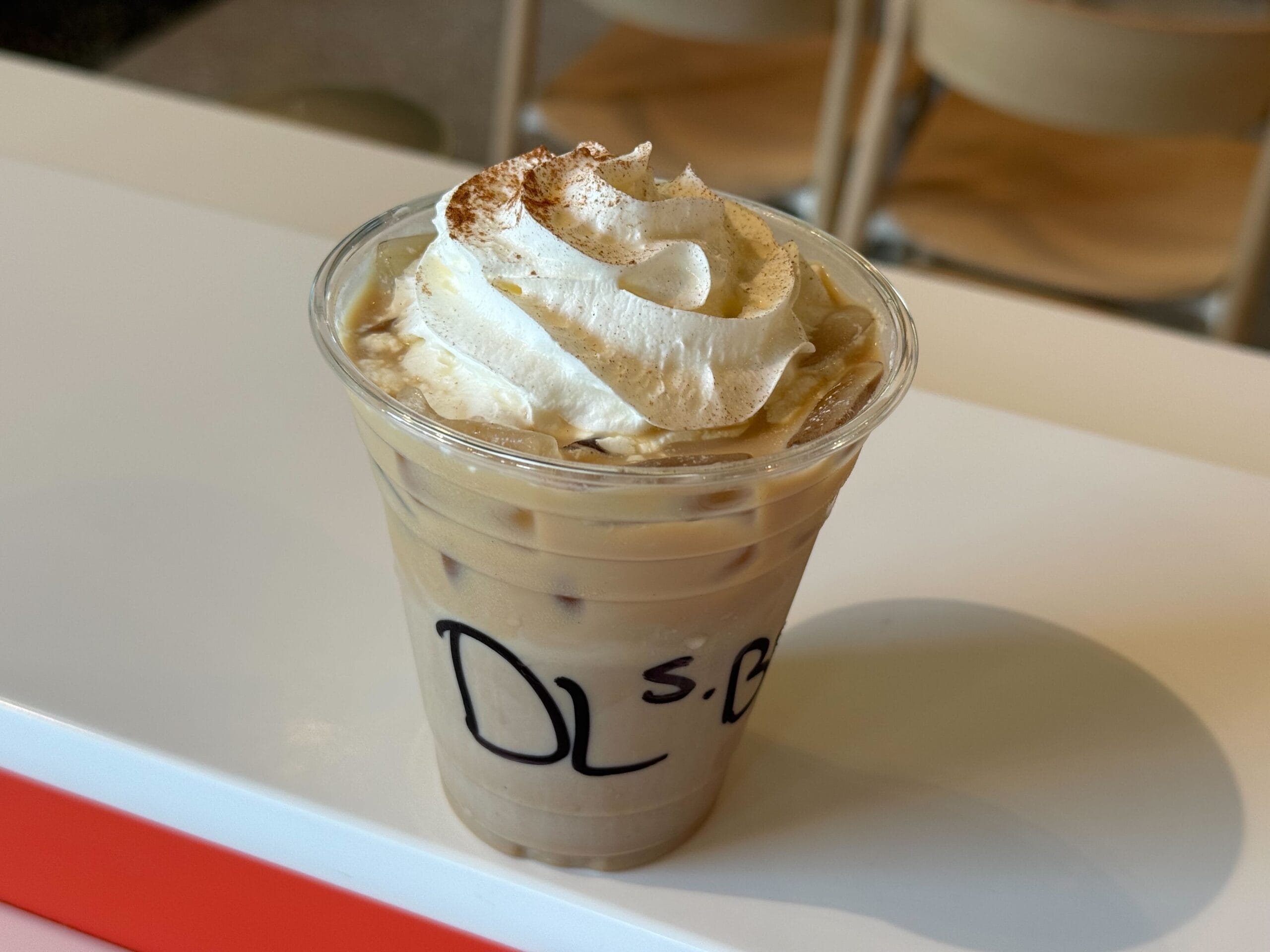 A plastic cup of iced coffee topped with whipped cream sits on a white table; a New Food & Wine treat from "DLS.B".