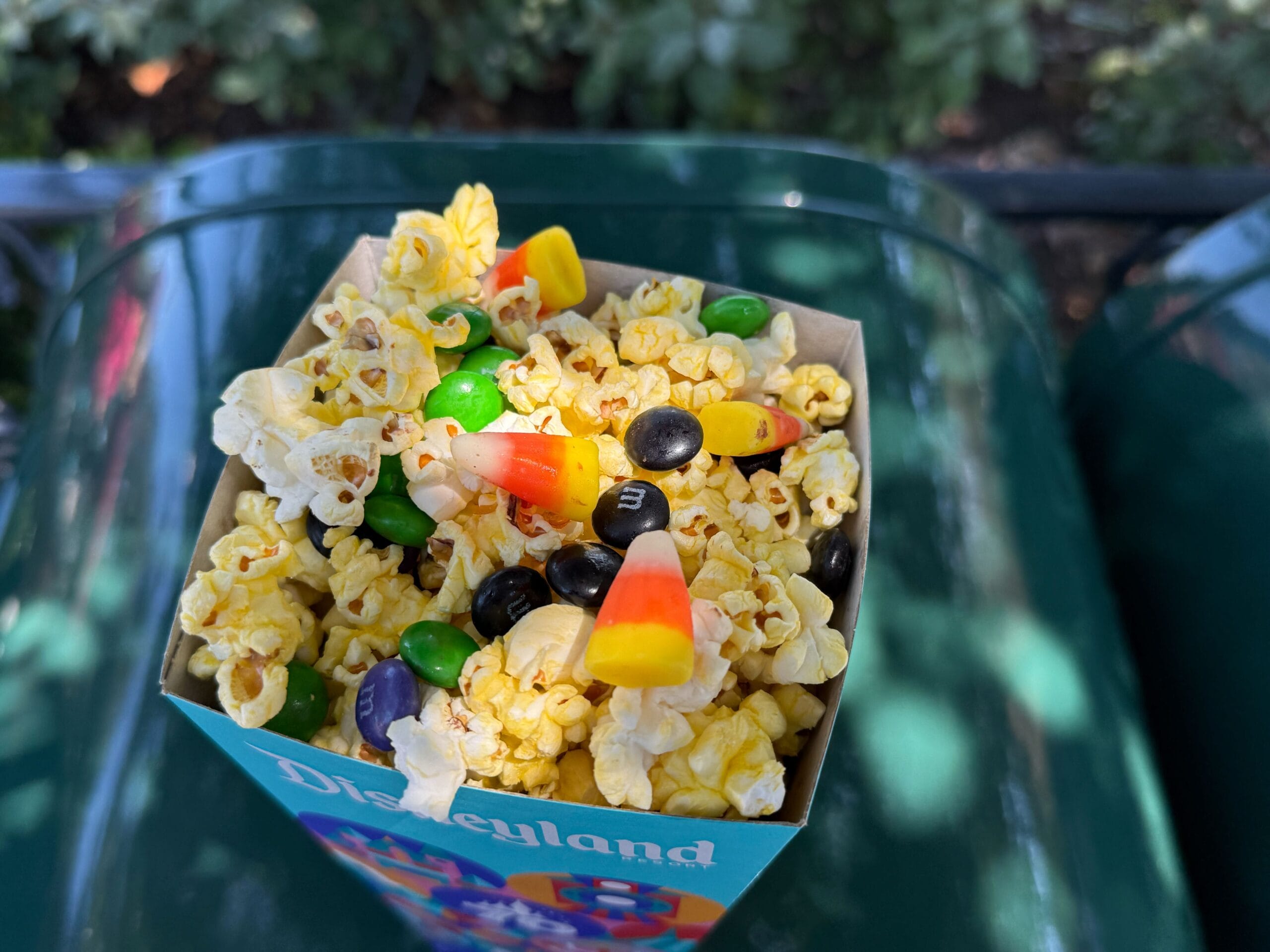 A Disneyland Halloween Treat Popcorn box topped with candy corn and chocolate candies sits on a green surface outdoors.