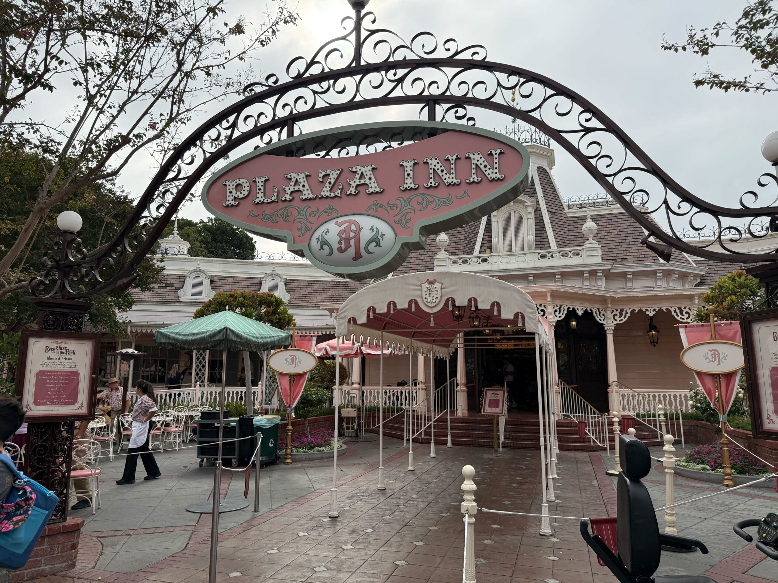 Entrance of Plaza Inn restaurant with ornate sign, pink decor, and outdoor seating area at Disneyland.