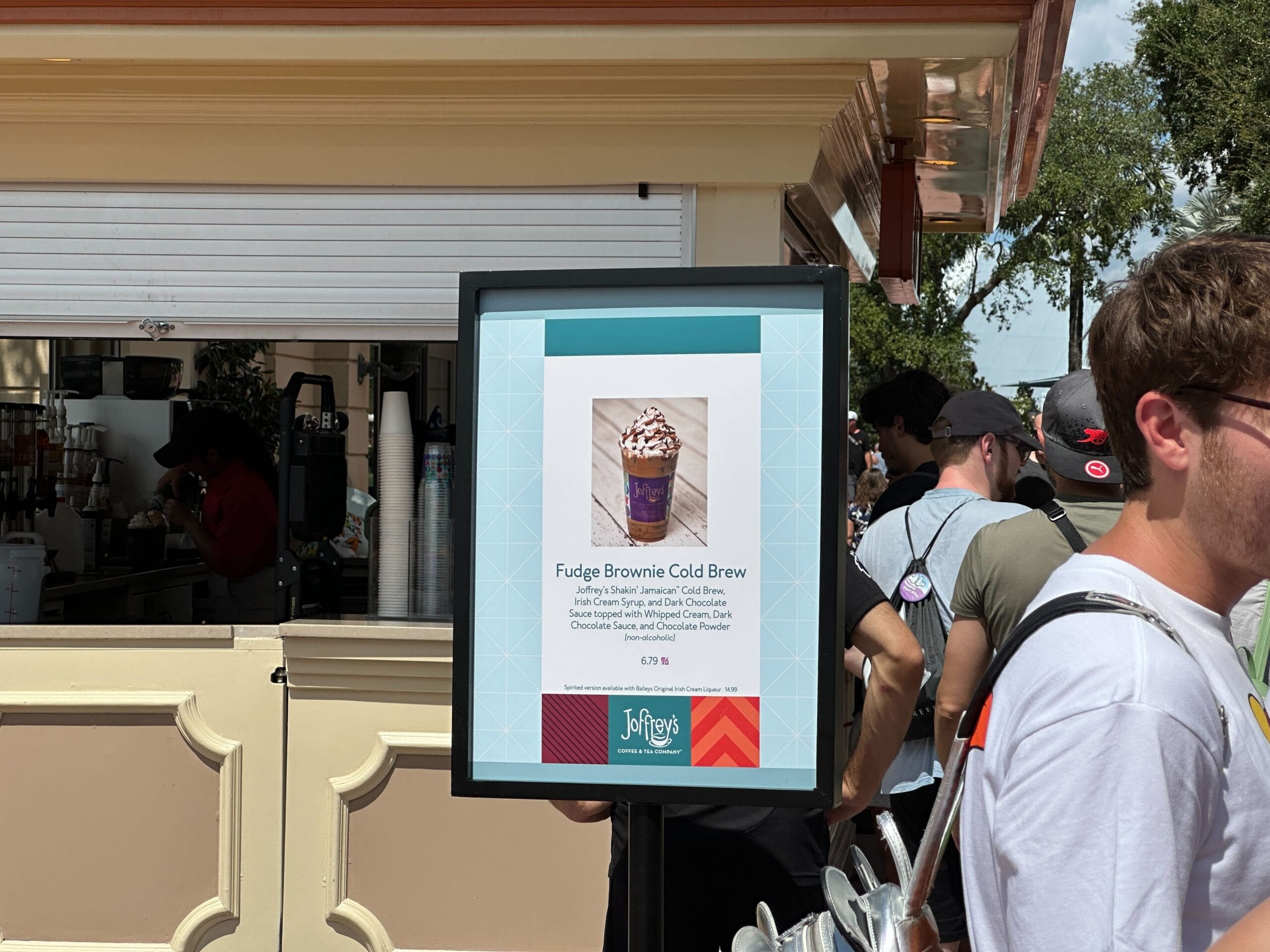 A sign displays a New Food & Wine Fudge Brownie Cold Brew outside a café, with people standing in line nearby.