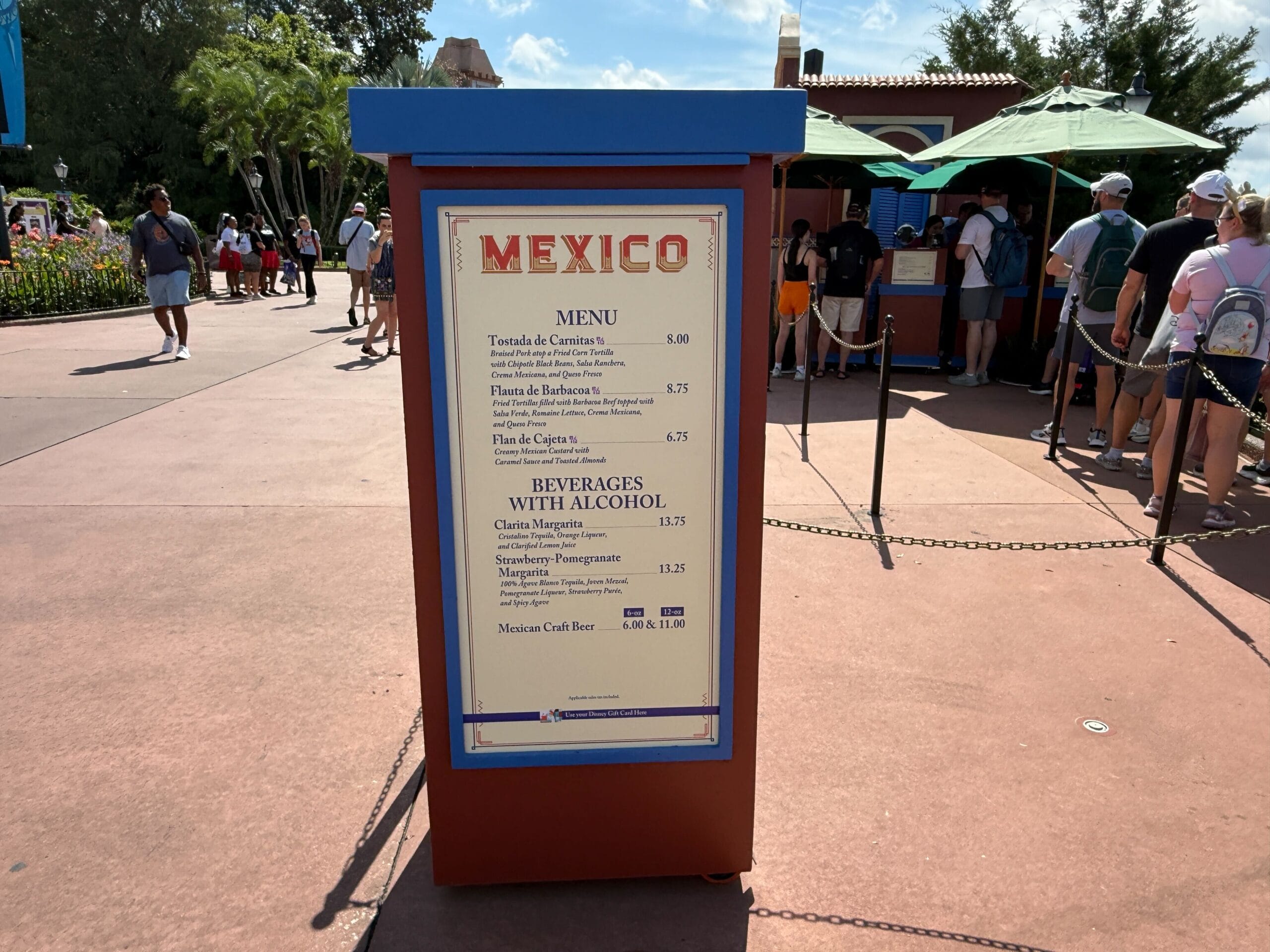 Menu board for Mexico pavilion at an outdoor New Food & Wine stand, with people in line and umbrellas in the background.