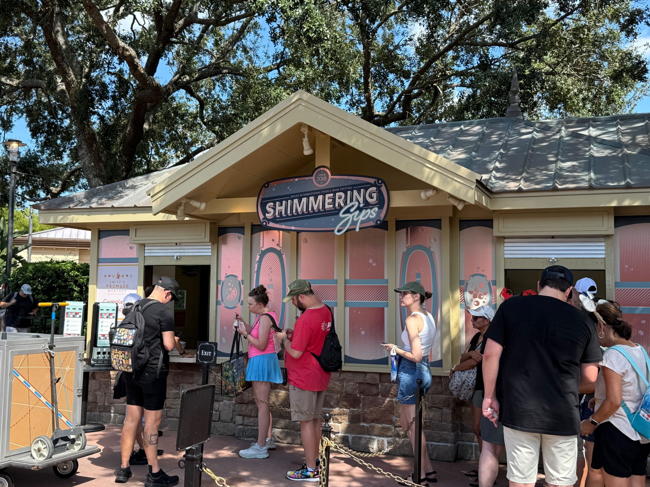 People stand in line at the Shimmering Sips kiosk during the Food & Wine festival under sunny trees.