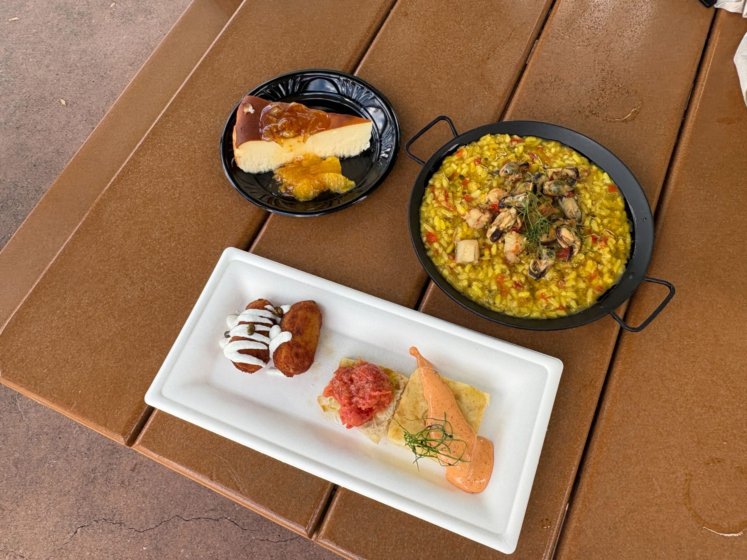 A wooden table with cheesecake, seafood rice, and Spanish tapas at the EPCOT Food and Wine Festival.