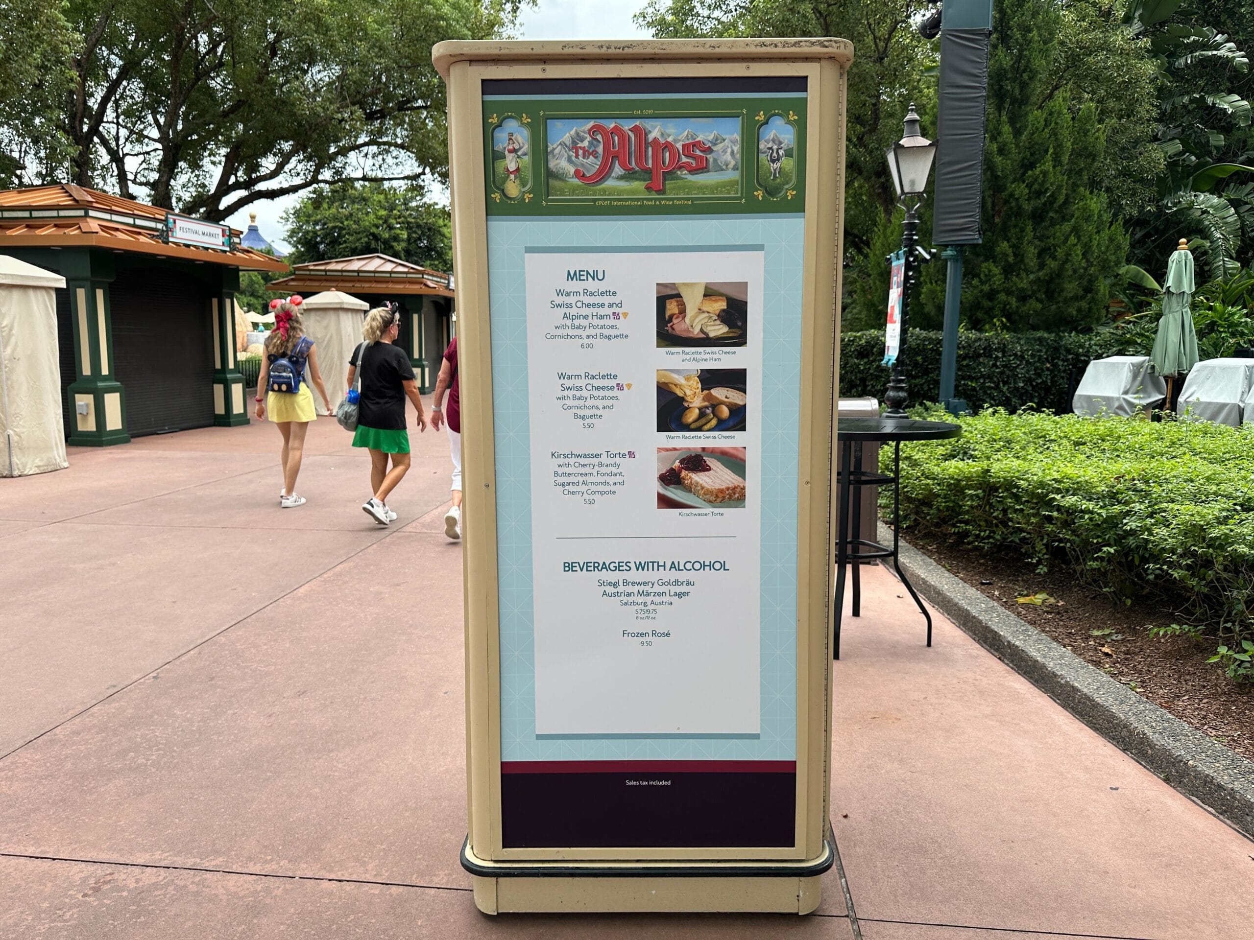 Outdoor menu display for The Alps, featuring Food & Wine options at an event, with people walking in the background.