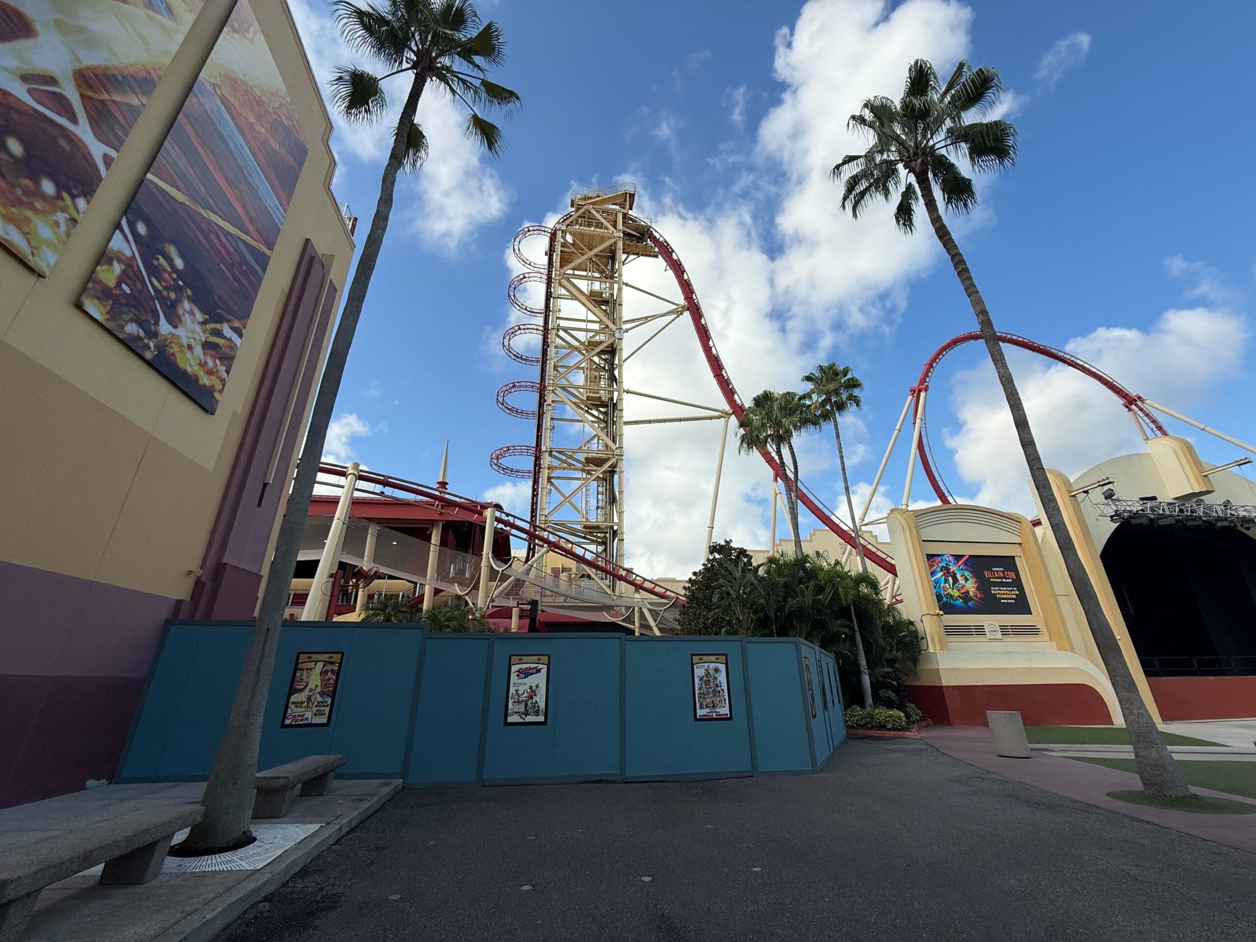 A tall roller coaster with red tracks and loops towers over palm trees and buildings under a partly cloudy sky.