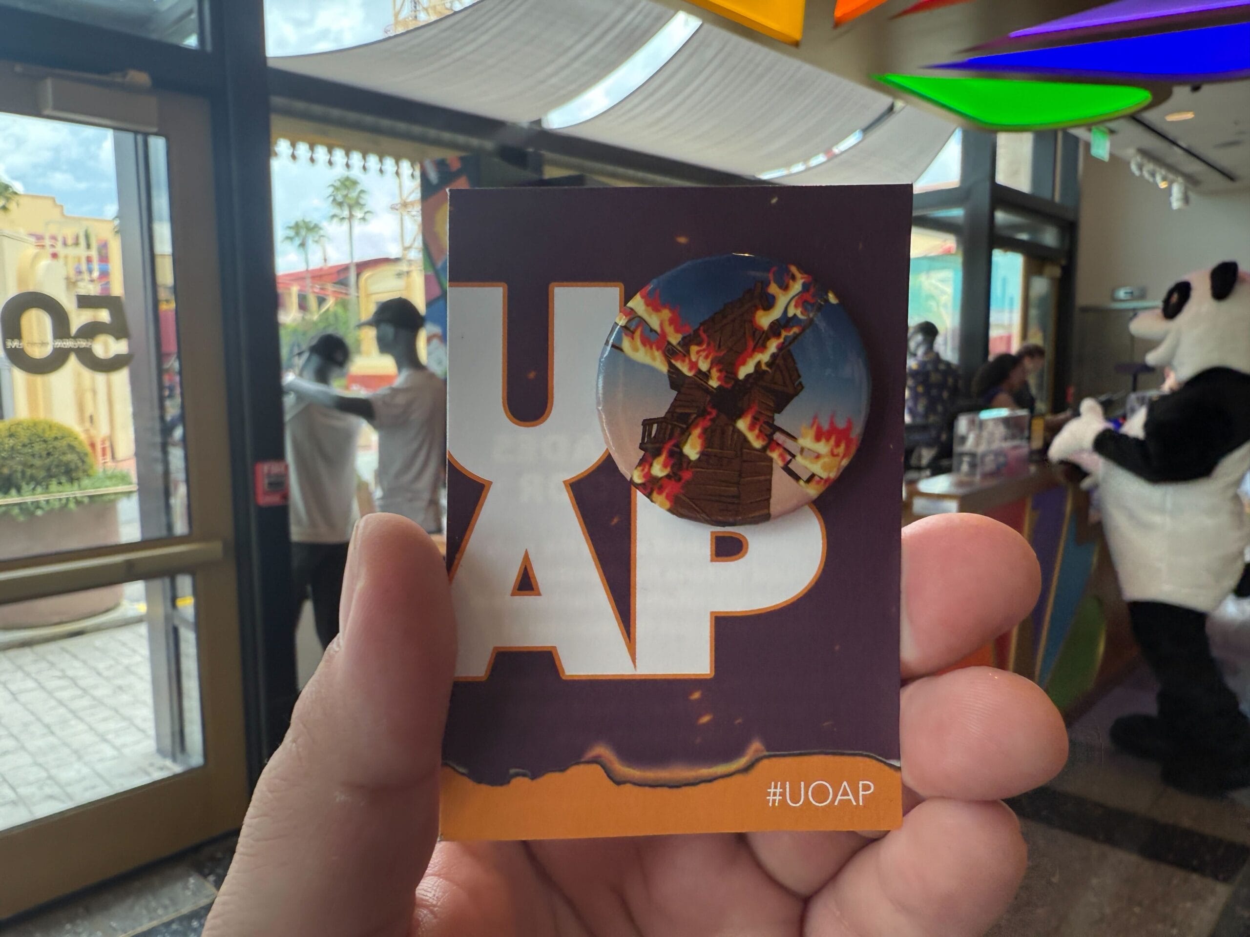 A hand holds a #UOAP card with a Burning Blade Tavern pin; people gather near a panda mascot at Universal Resort.