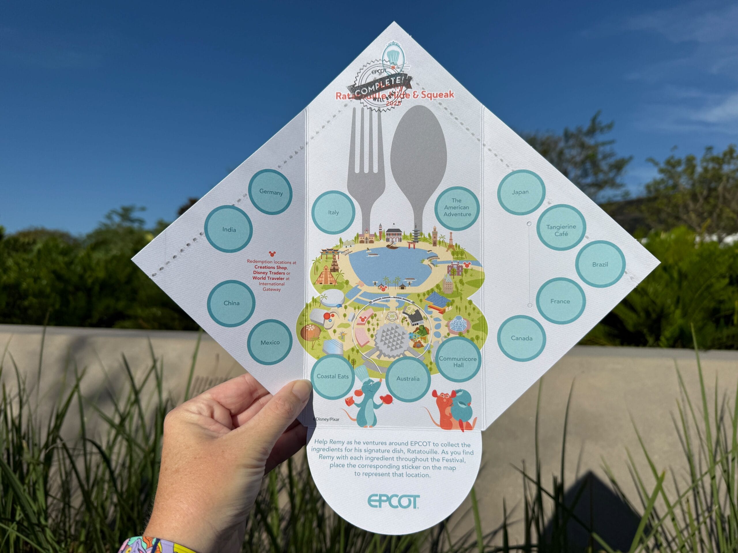 A hand holds a colorful EPCOT-themed scavenger hunt map outdoors, featuring country names and park illustrations.