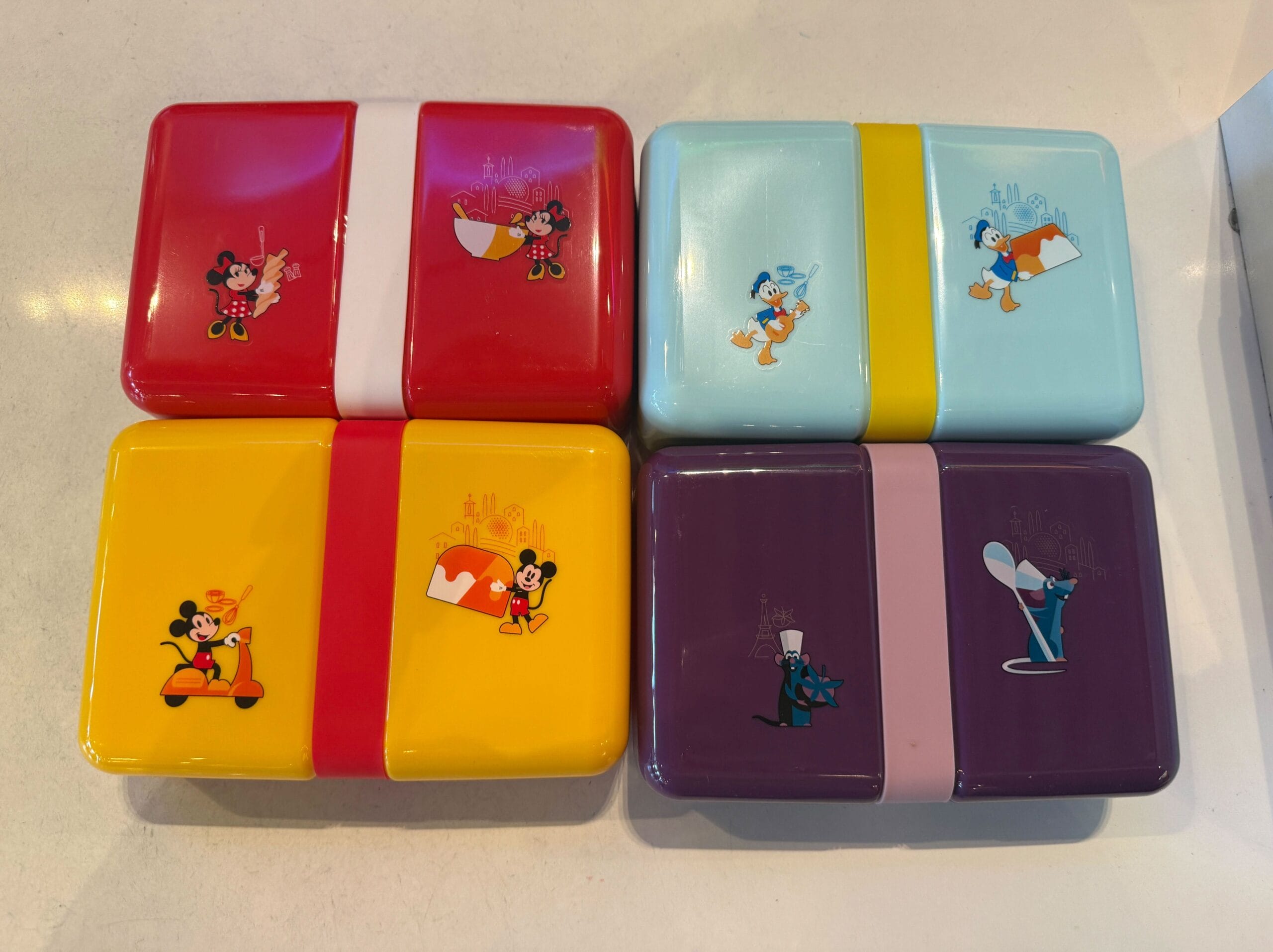 Four rectangular Disney lunchboxes in red, blue, yellow, and purple—perfect for your next EPCOT Food & Wine Festival adventure.
