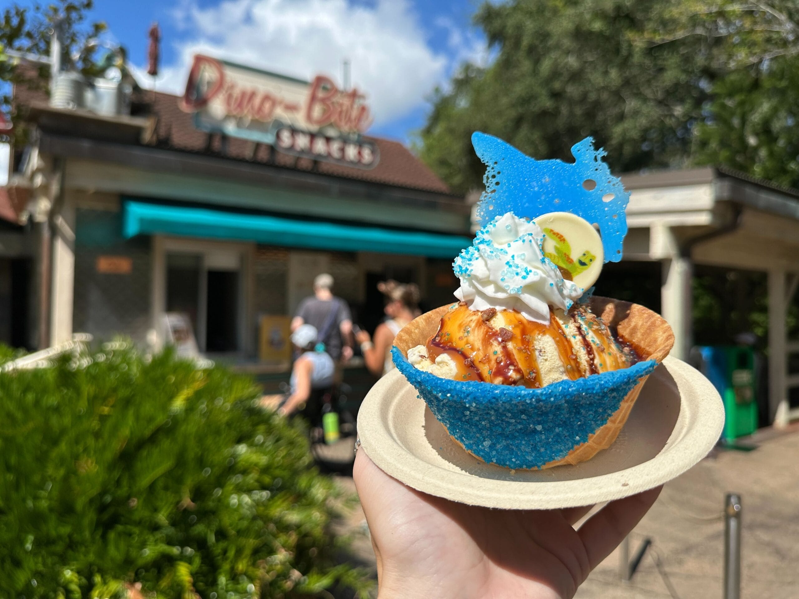 A hand holds a Totally Sweet Sundae in front of Dino-Bite Snacks, topped with blue sugar and white chocolate.
