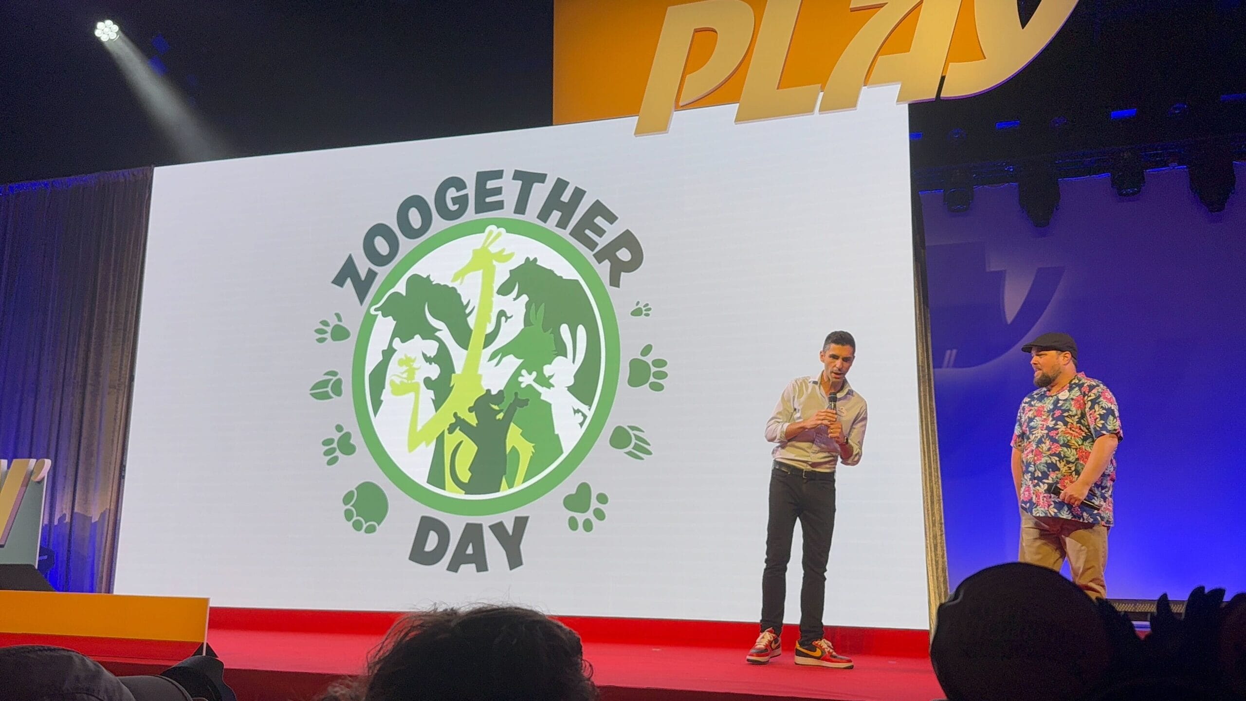 Two people stand on stage by a screen showing the "Zoogether Day" logo, featuring Zootopia: Better Zoogether animal silhouettes.