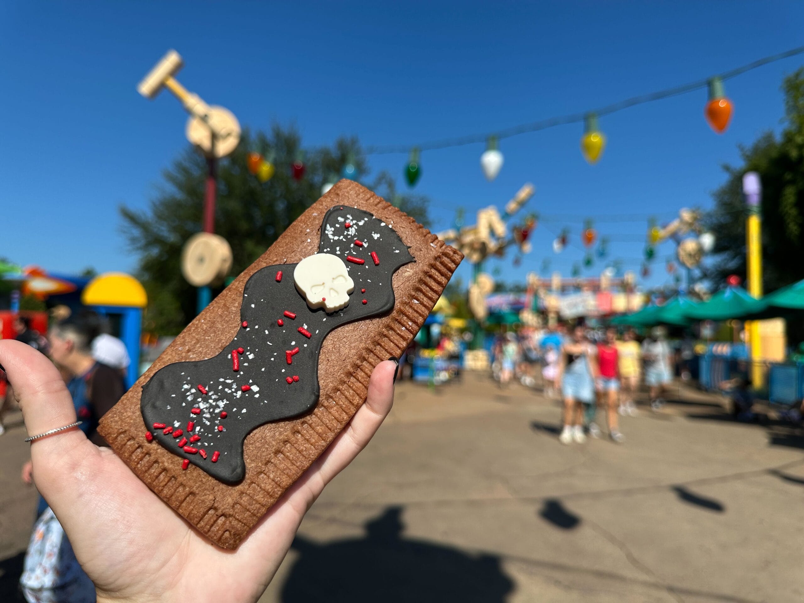 A hand holds a Spiced Berry Lunch Box Tart at Hollywood Studios, surrounded by festive decor and a lively crowd.
