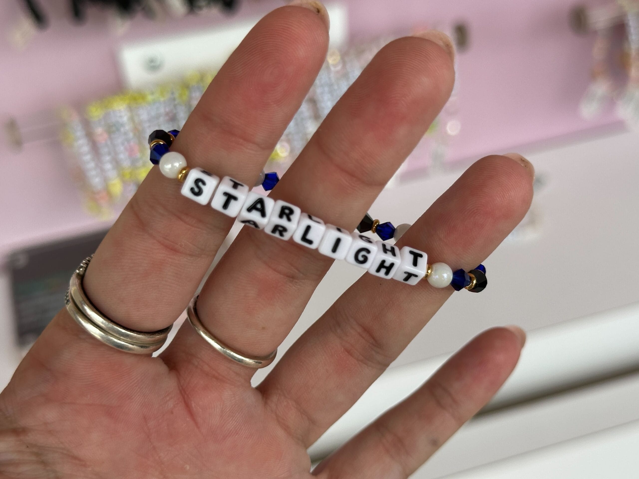 A hand holds a Little Words Project Starlight bracelet with white letter beads spelling “STARLIGHT.”