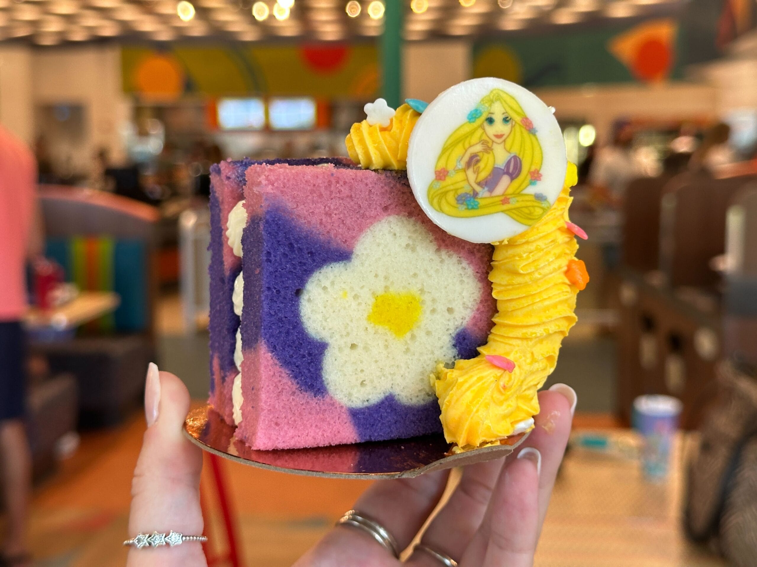 A hand holds the Rapunzel Berries and Cream Cake at Everything POP Shopping & Dining, topped with a white chocolate Rapunzel.