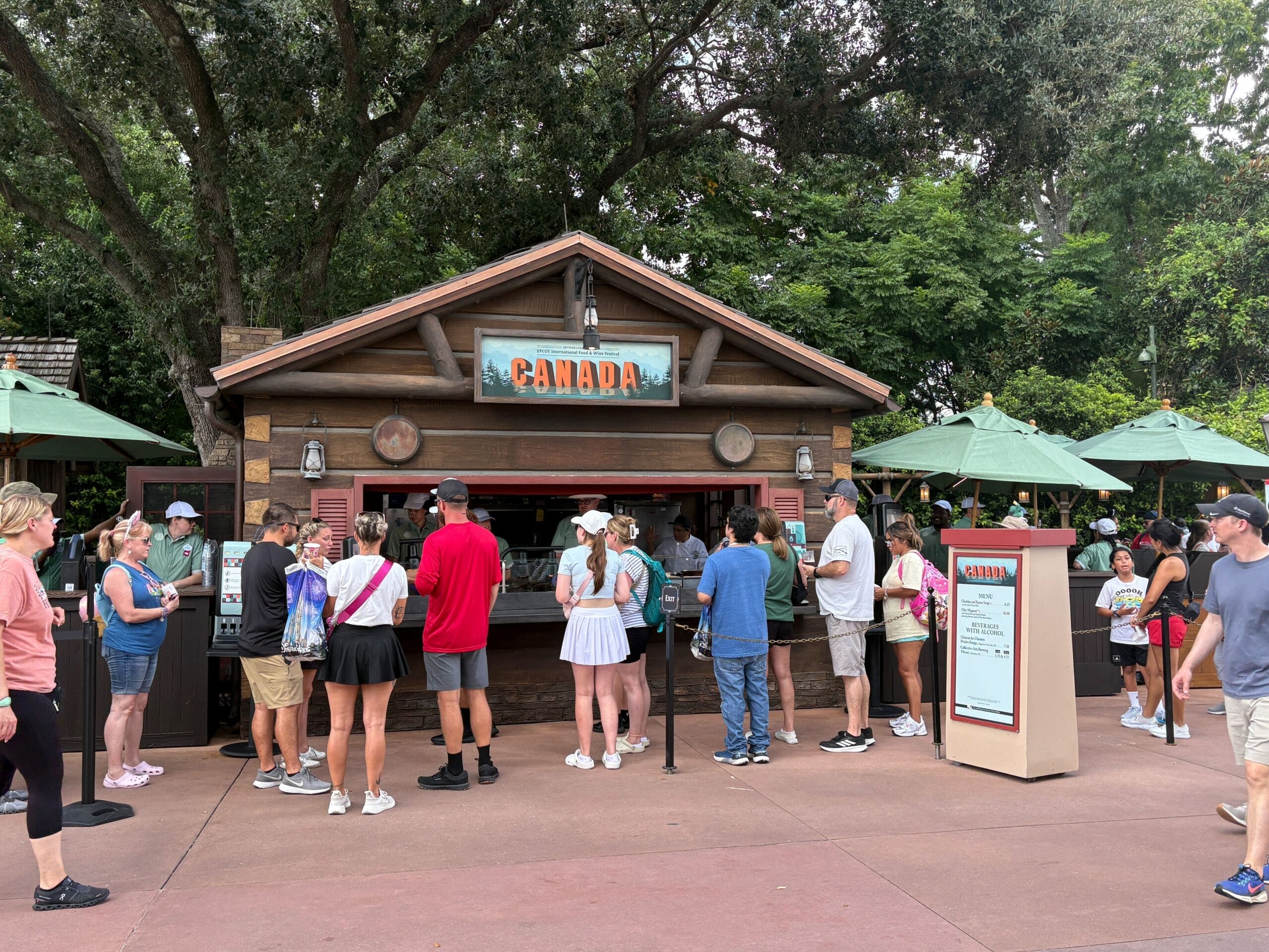 People line up at the EPCOT 2025 Food and Wine Festival’s Canada booth, surrounded by trees and green umbrellas.