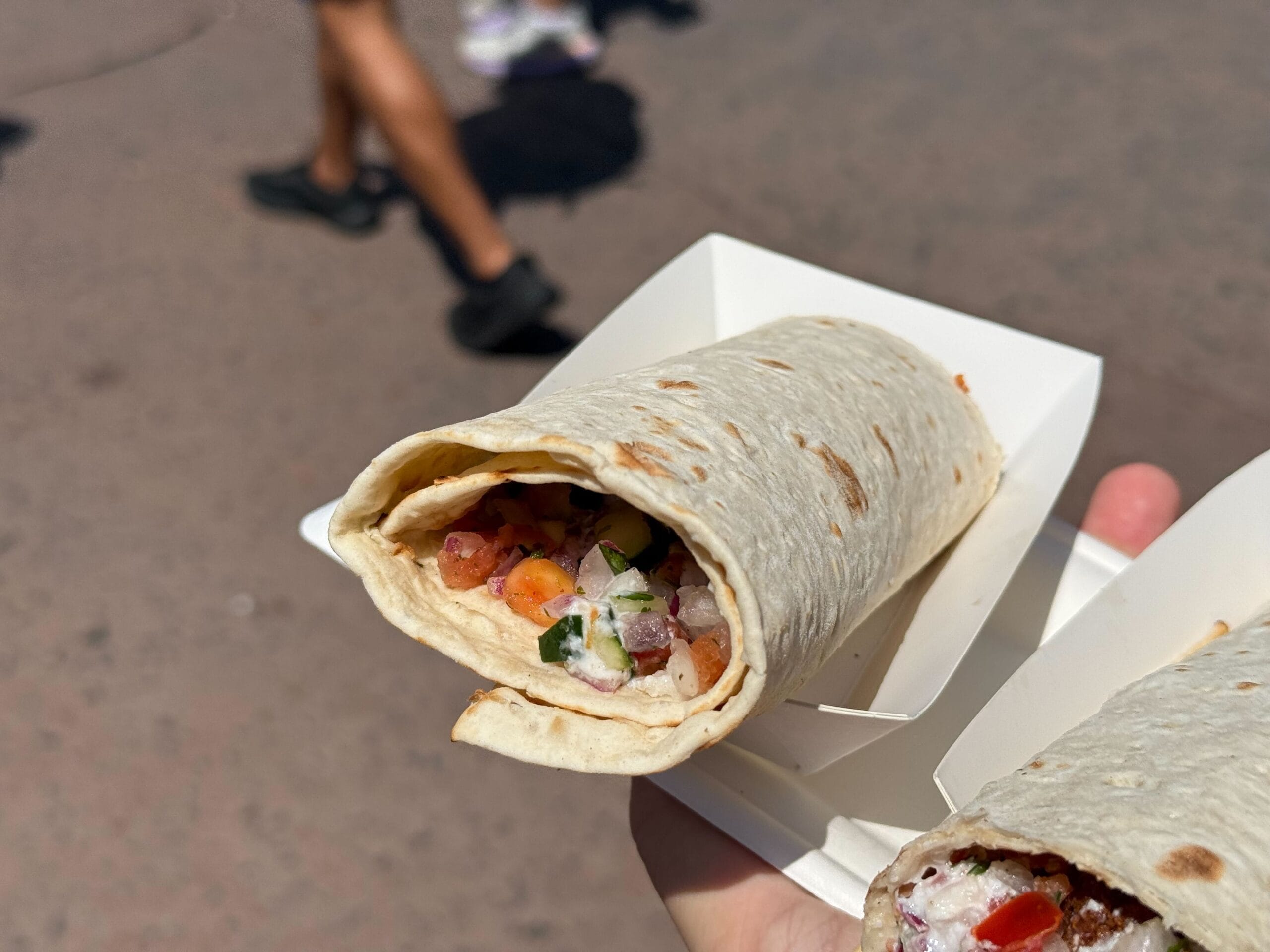 A hand holds a Food & Wine tortilla wrap filled with diced vegetables in a white paper tray; people walk in the background.