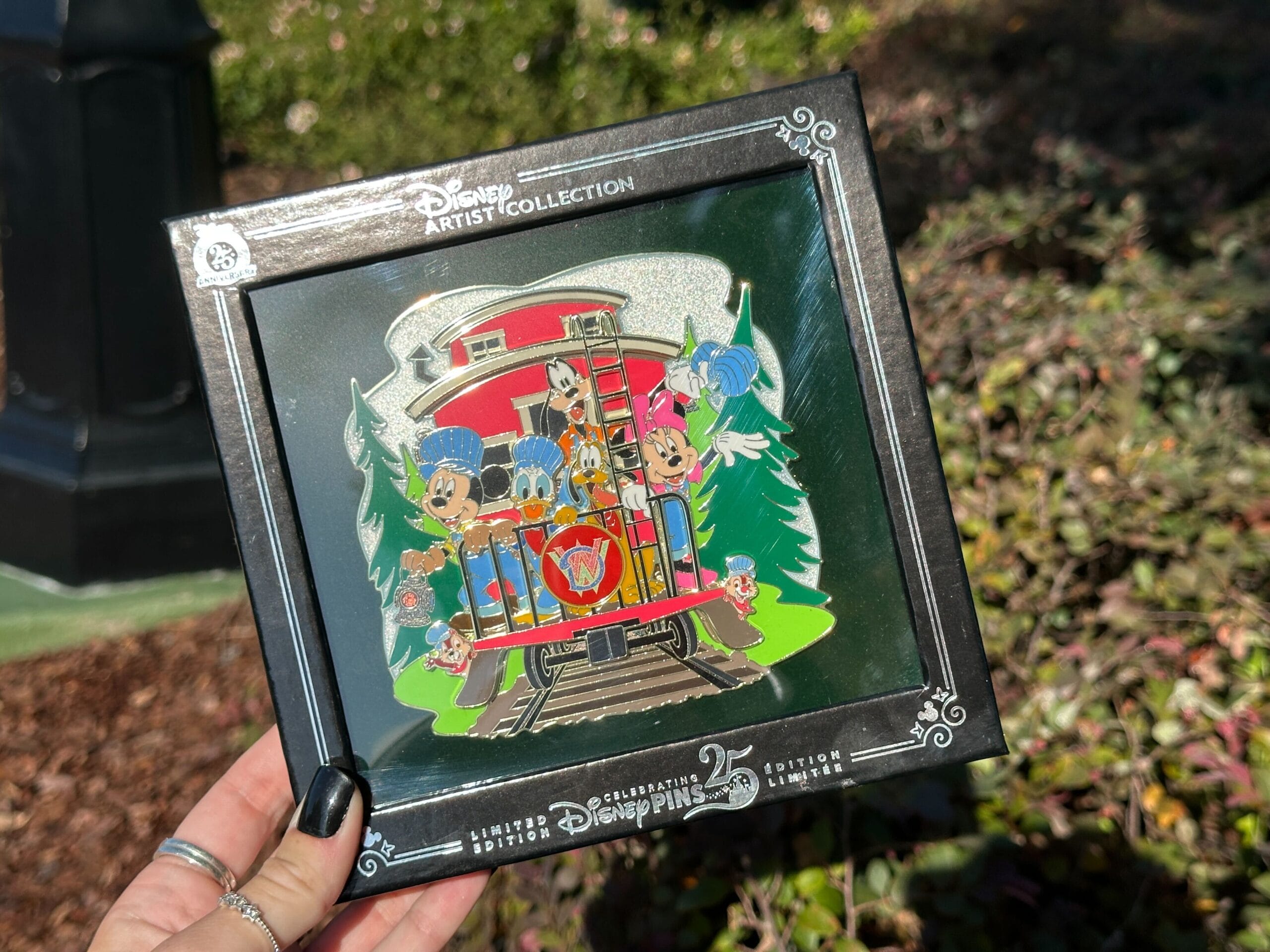 A hand holds a Disney Artist Collection pin featuring Mickey, Minnie, and friends on a red trolley surrounded by trees.