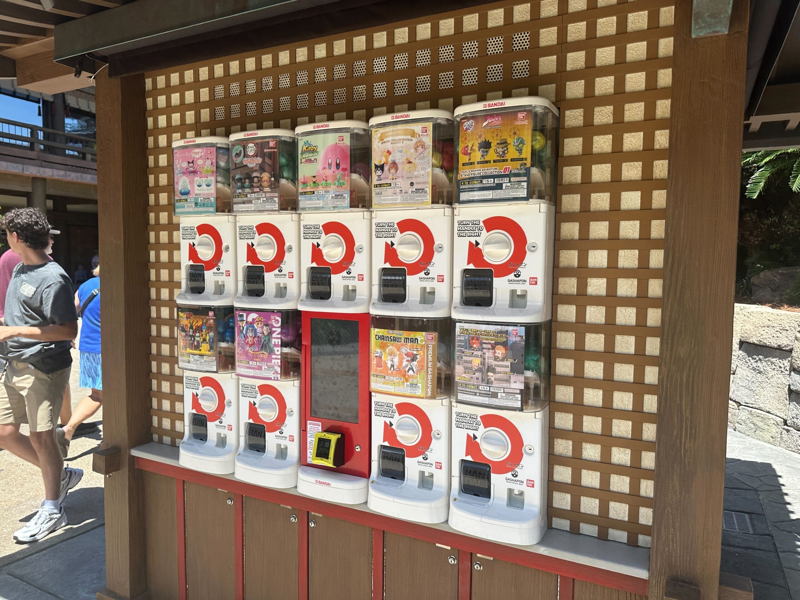 A row of Japanese capsule toy vending machines, called gachapon, displayed outdoors against a wooden lattice wall.