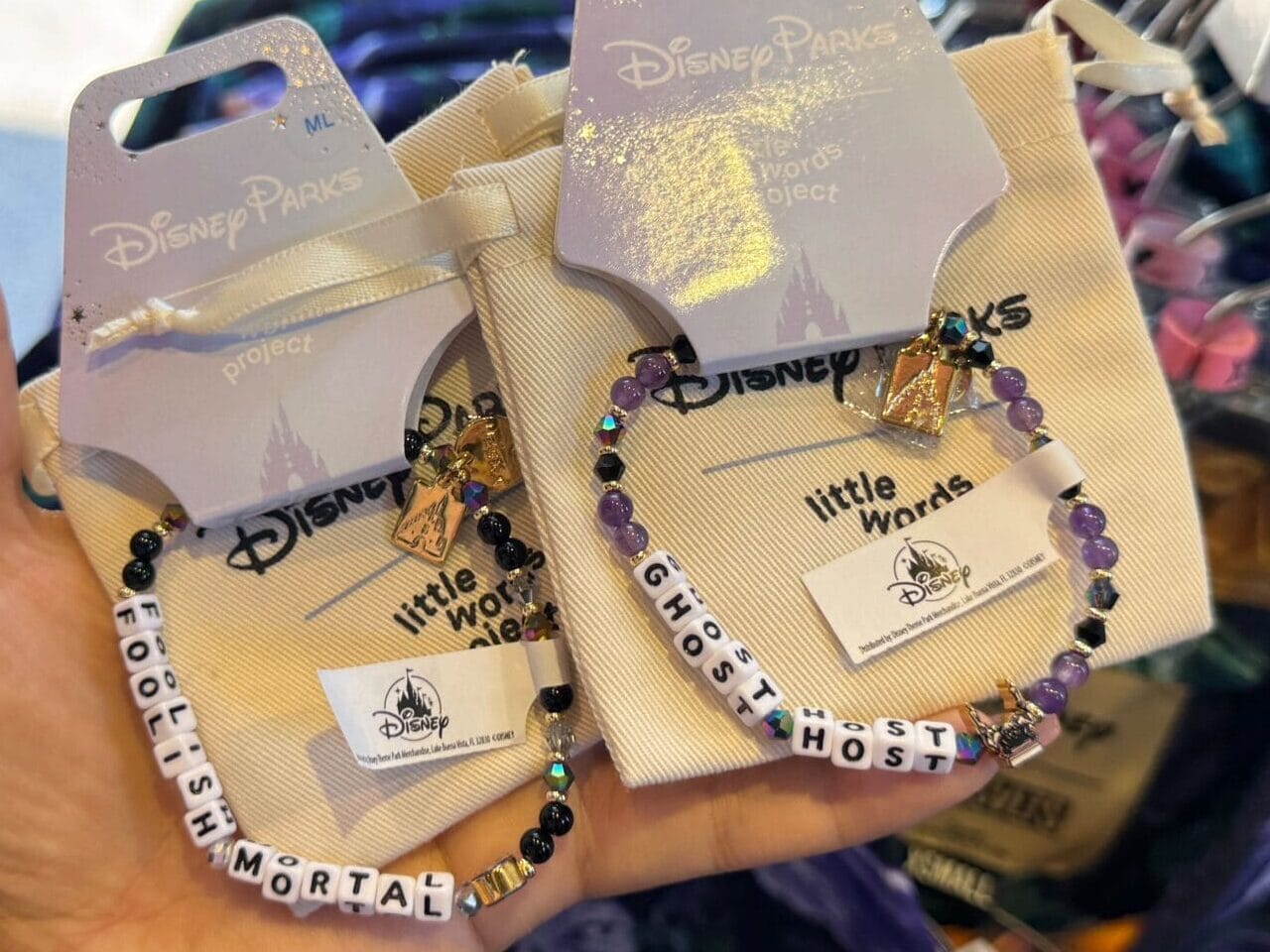 A hand holds two "little mortal" bracelets at a Disneyland Haunted Mansion merchandise display.