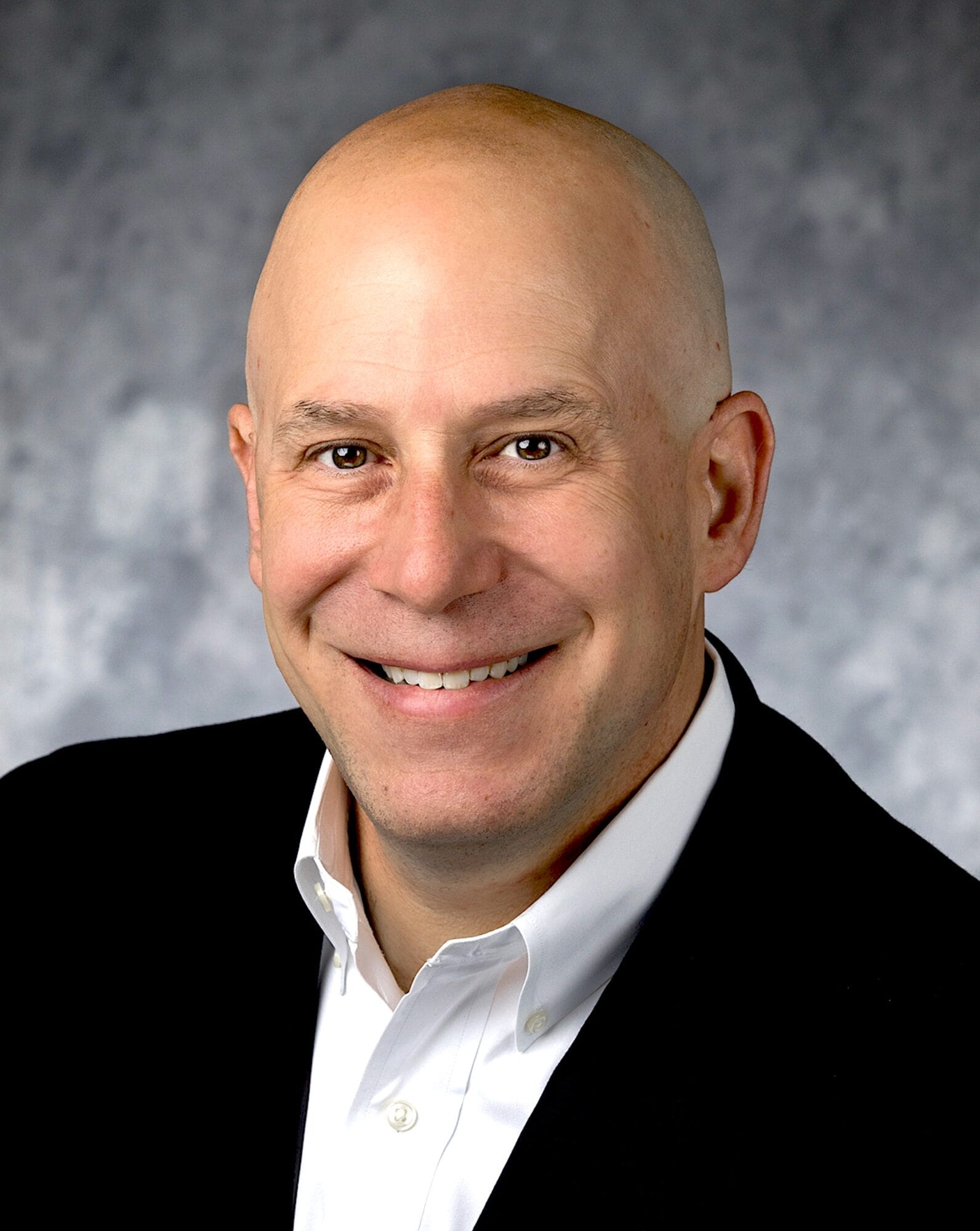 Smiling bald man in a black suit and white shirt, Creative Consultant, against a gray, textured background.