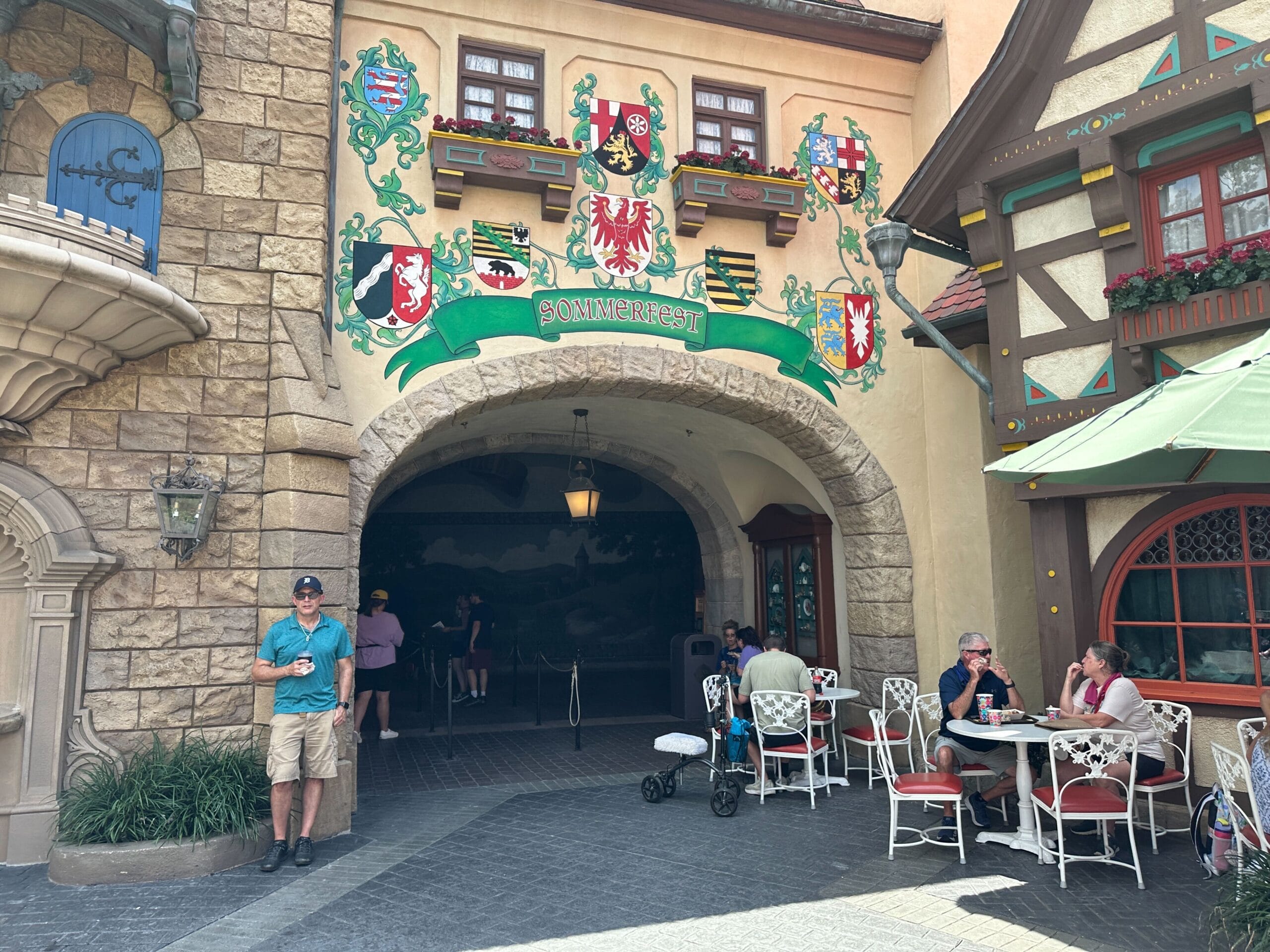 Man stands near stone building with decorated archway; Sommerfest guests sit at outdoor tables under umbrellas.