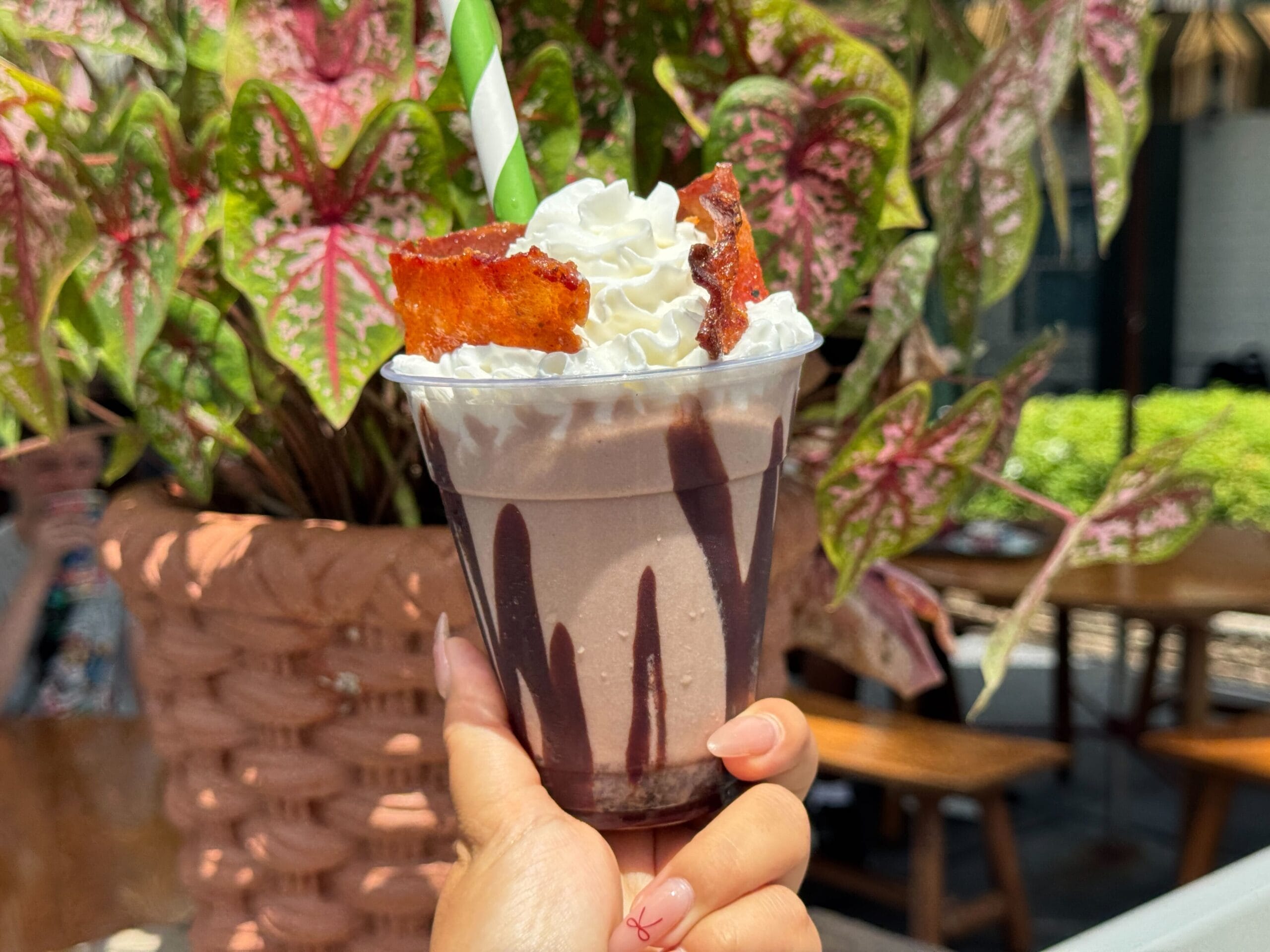 A hand holds a Chocolate Bourbon Milk Shake with whipped cream and bacon pieces in front of leafy plants outdoors.