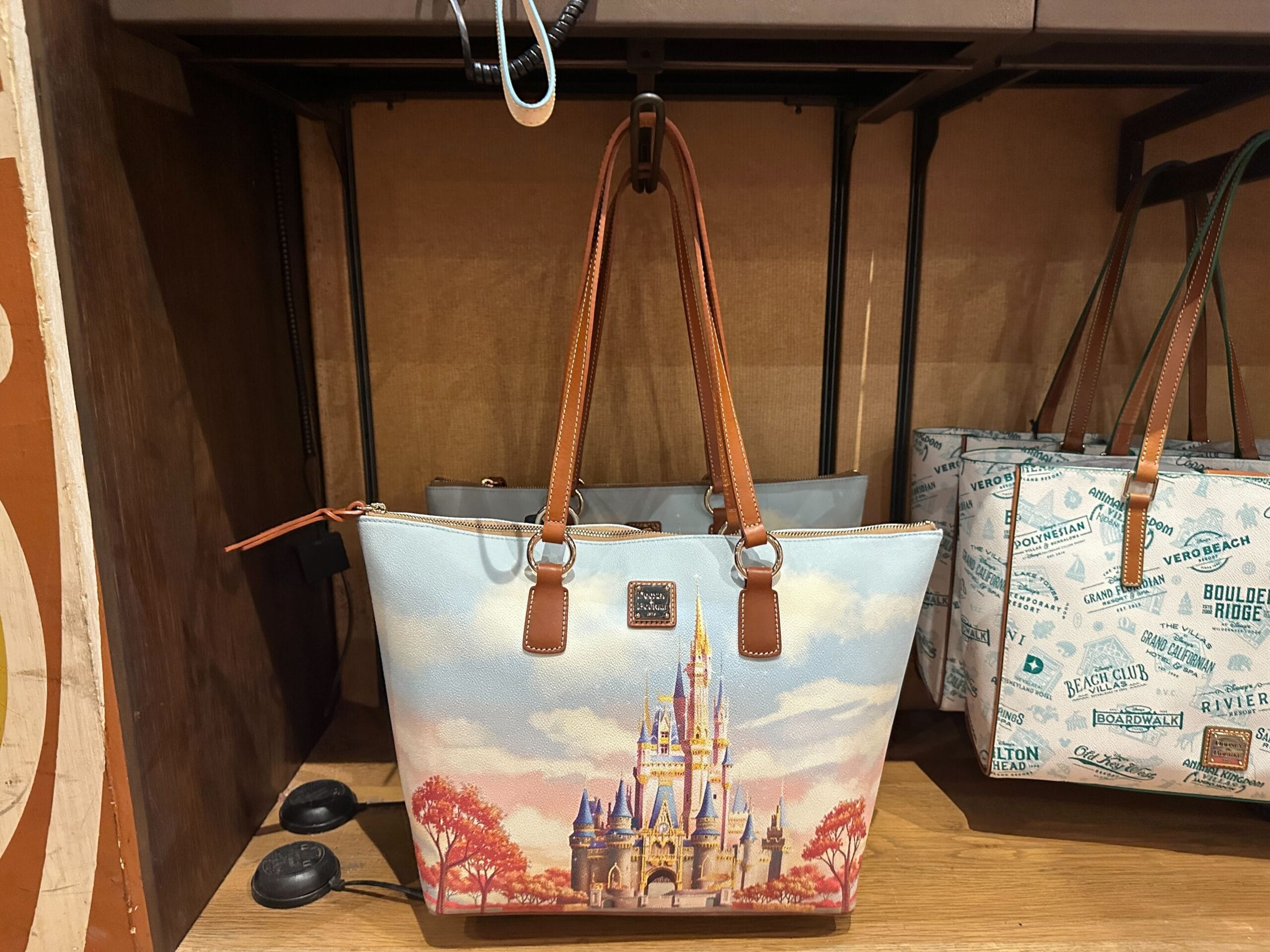 A handbag with a Cinderella Castle design and brown handles is displayed on a wooden shelf.