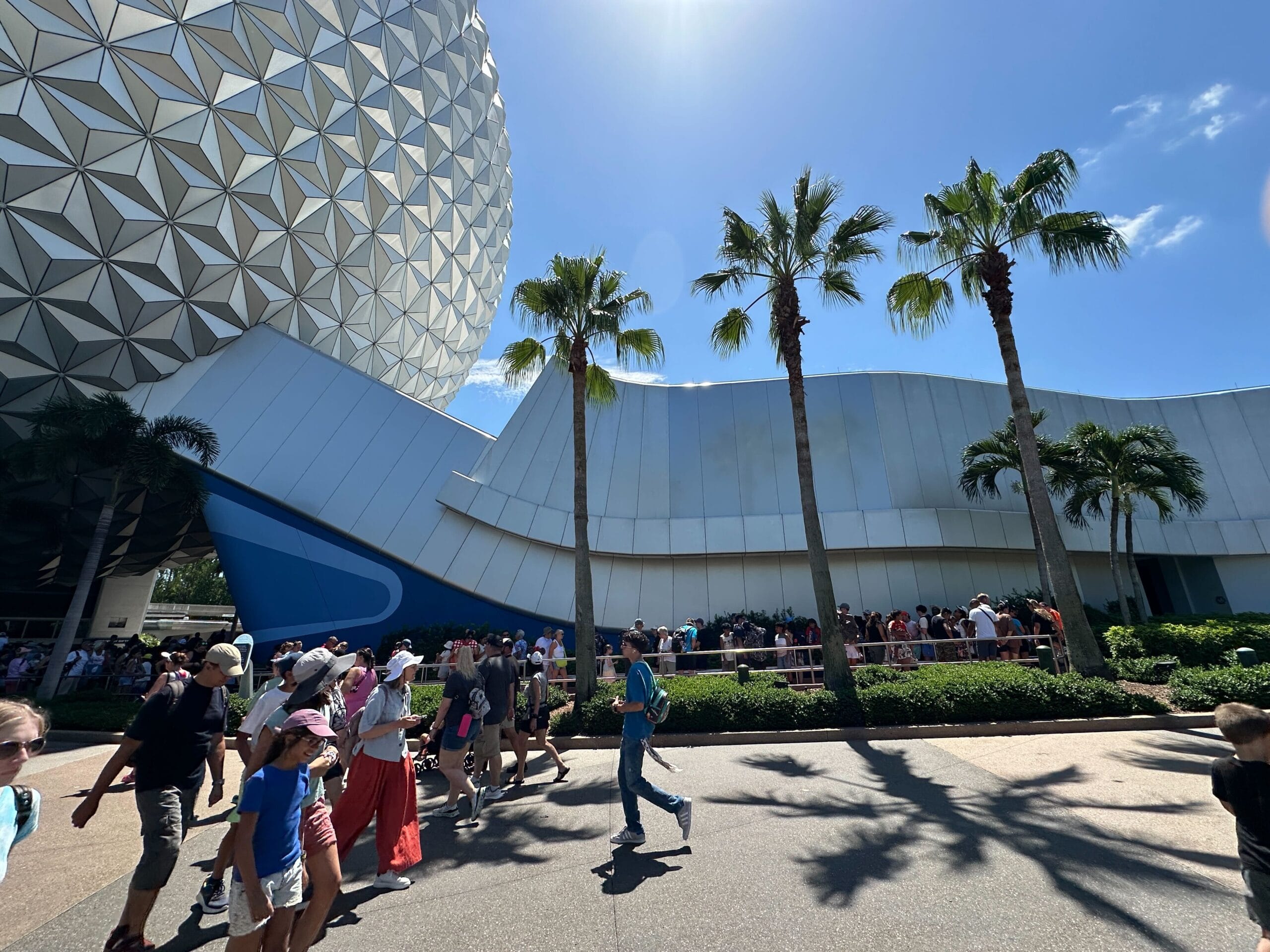 Crowds walk past palm trees and the geometric dome at Epcot on a sunny day, with people lined up near the building.