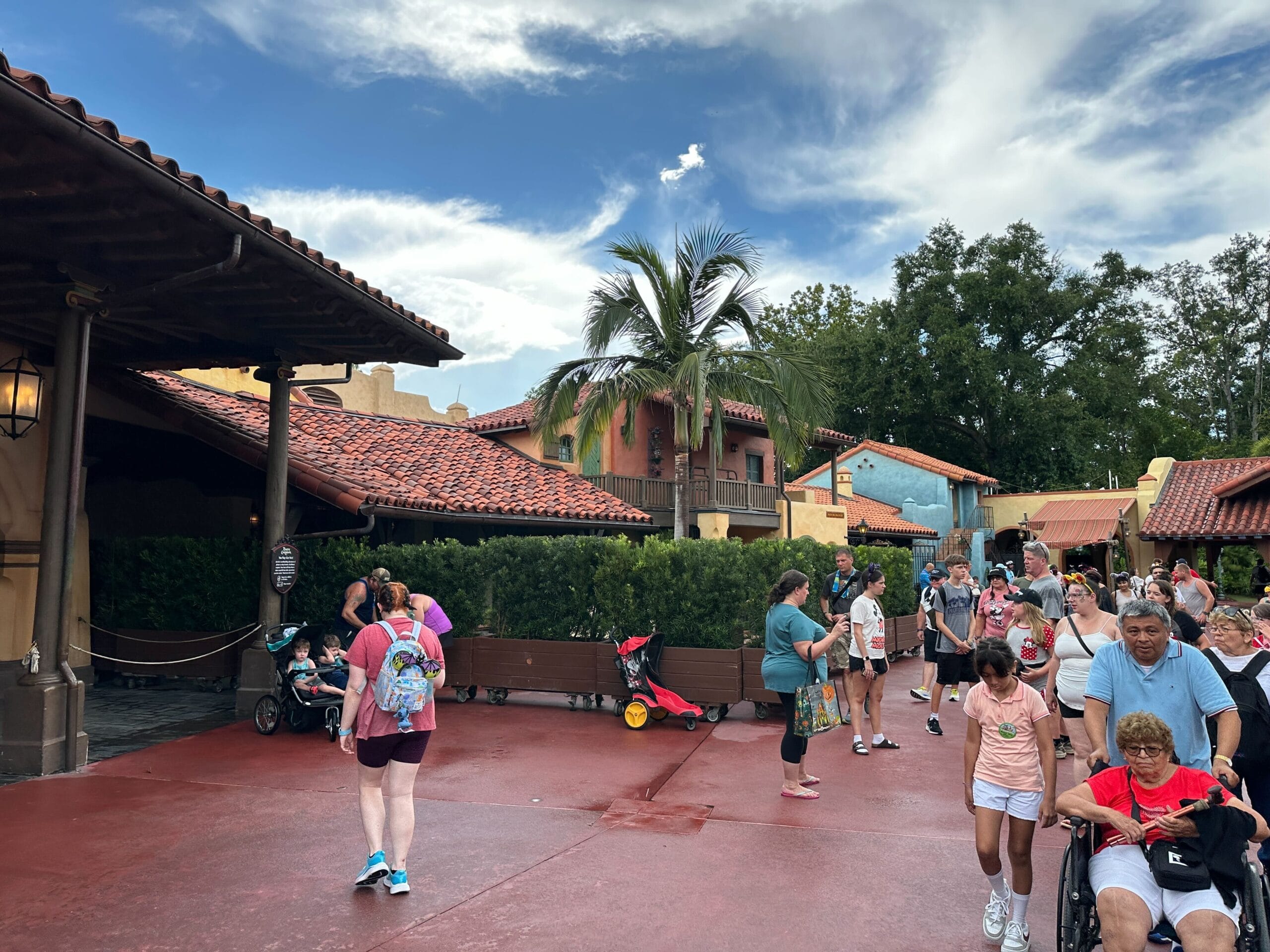 People walk along a path with colorful buildings, palm trees, and benches under a partly cloudy sky.