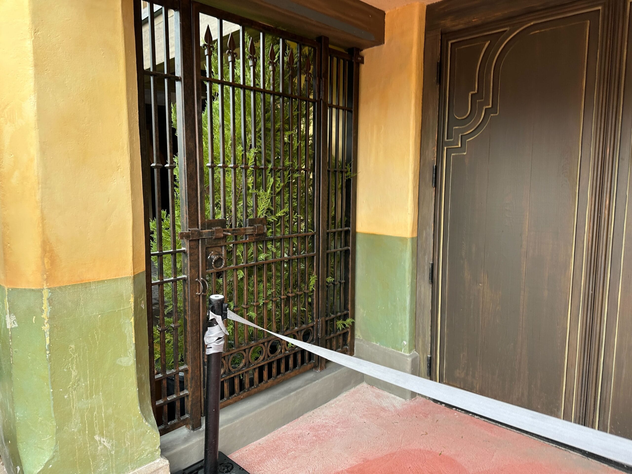 A locked metal gate by a brown door, with construction walls in front and green plants behind the barrier.
