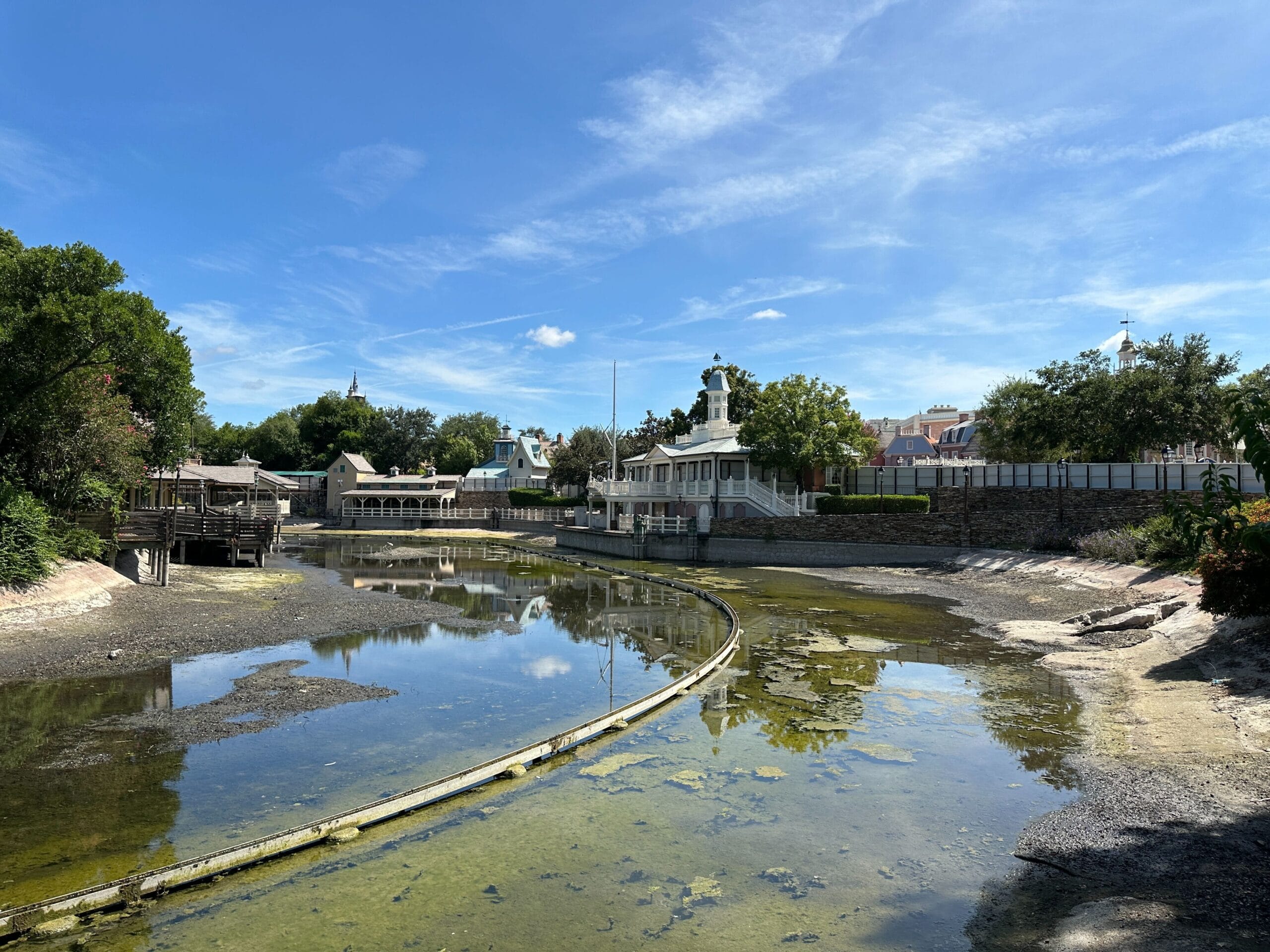 A shallow, partially drained river with buildings and trees under a bright blue sky with wispy clouds.