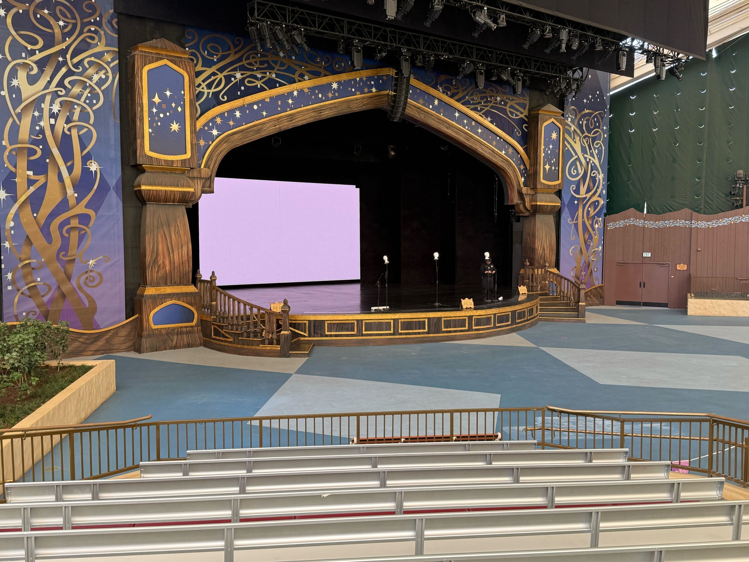 Empty outdoor stage at Fantasyland Theatre, adorned in gold and purple, viewed from empty seating at Disneyland.
