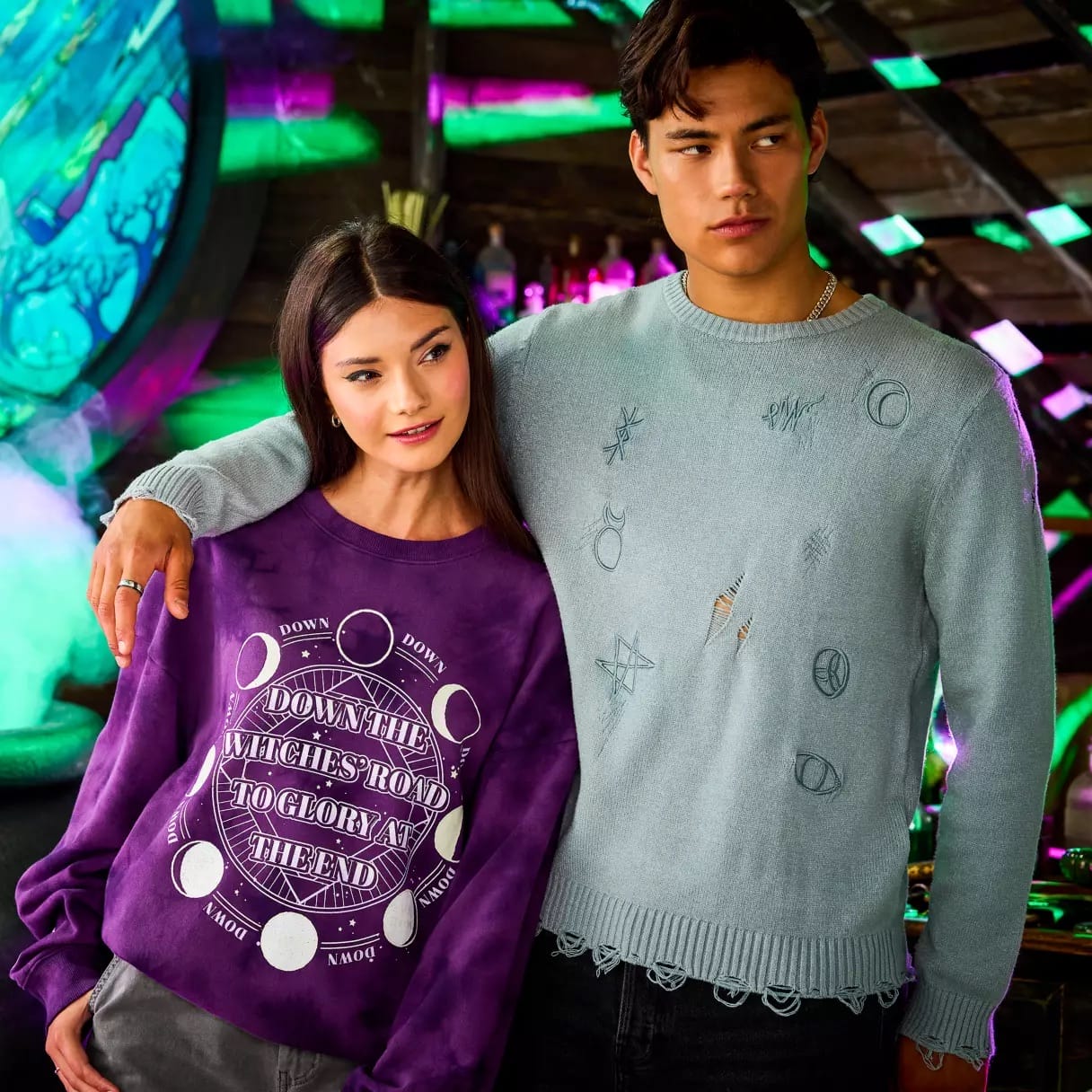 A woman in a purple Agatha All Along sweater stands next to a man in new Disney Store apparel with celestial symbols.