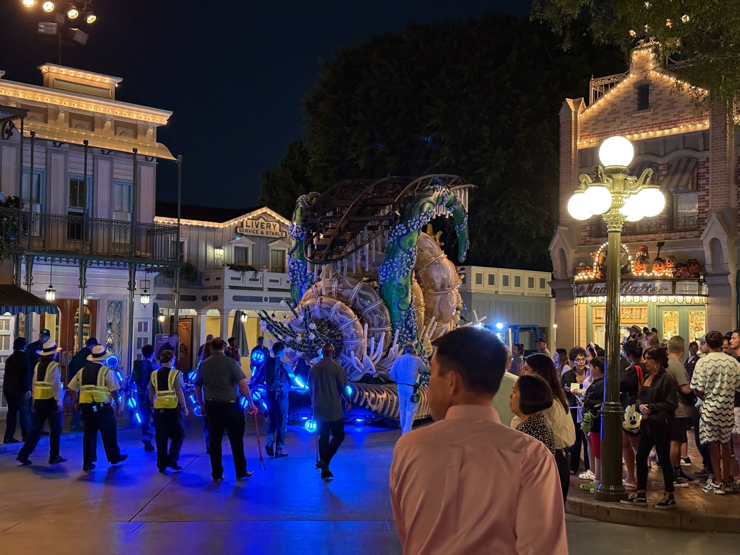 A night parade with a large dragon float and crowds watching on a lit-up street, with staff in yellow vests nearby.