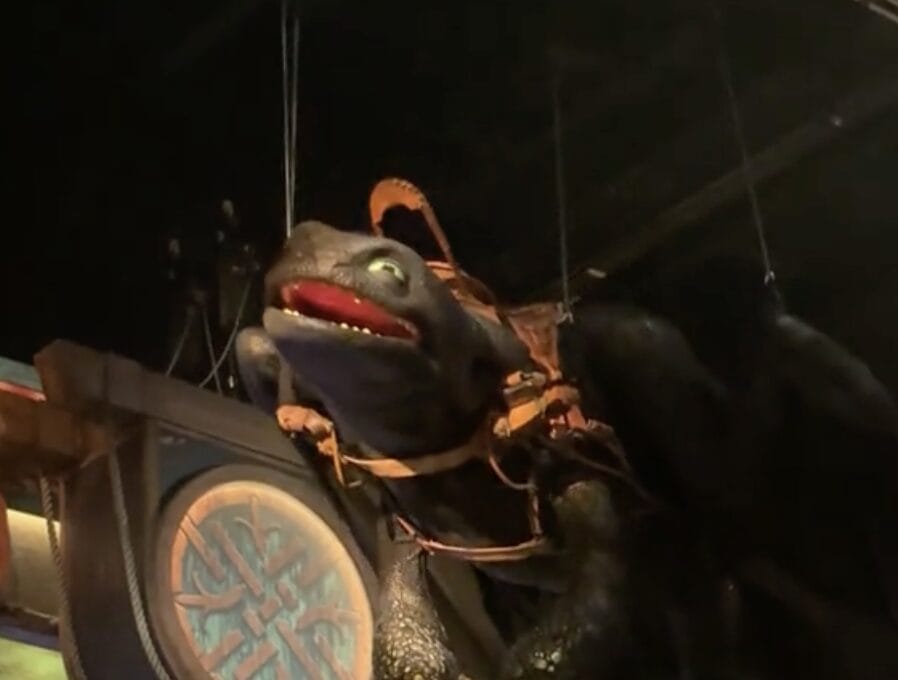 A large black Toothless dragon animatronic with green eyes and a saddle is suspended indoors by wires.
