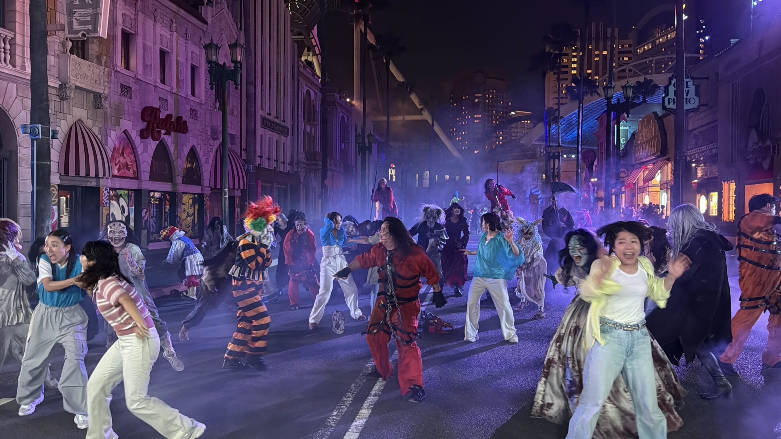 A group in spooky costumes performs Zombie de Dance on a foggy city street under colorful lights.