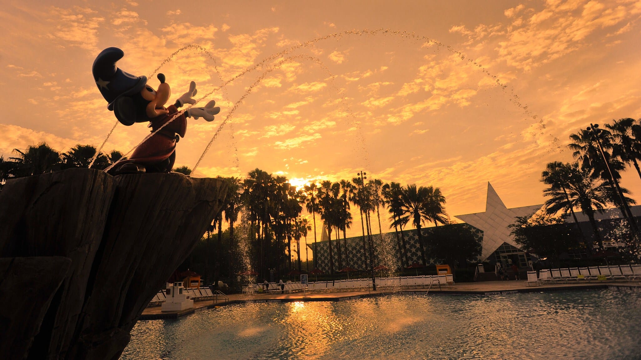Statue of Sorcerer Mickey sprays water at Fantasia Kiddie Pool, Disney's All-Star Movies Resort, with sunset and palm trees.