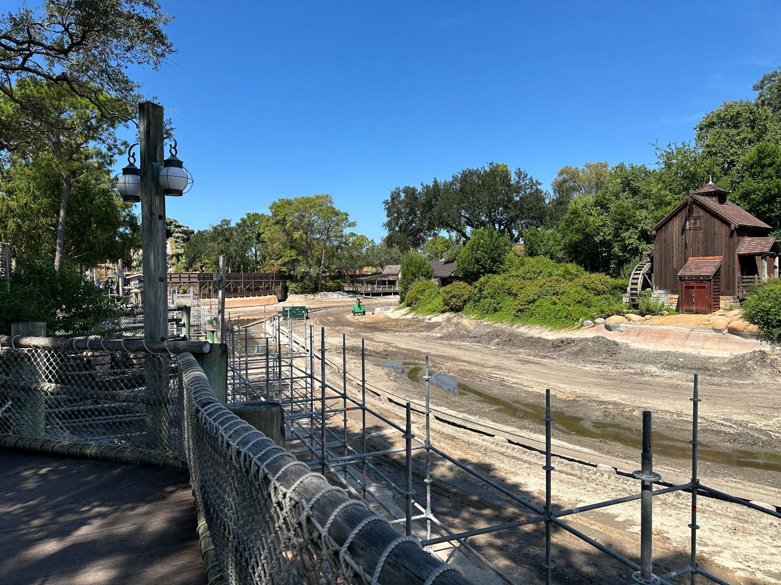A construction update shows a dry Rivers of America riverbed with scaffolding, trees, and rustic buildings under a clear blue sky.