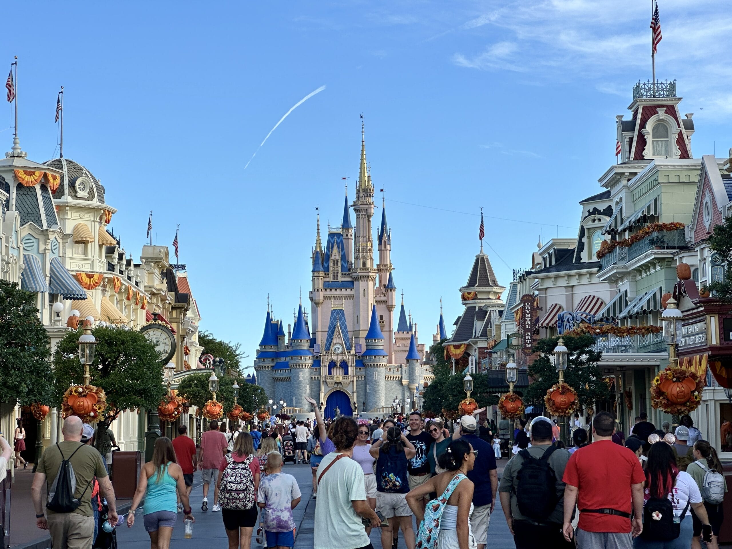 Crowds walk down Main Street toward the Magic Kingdom castle, with fall decor and a clear blue sky above.