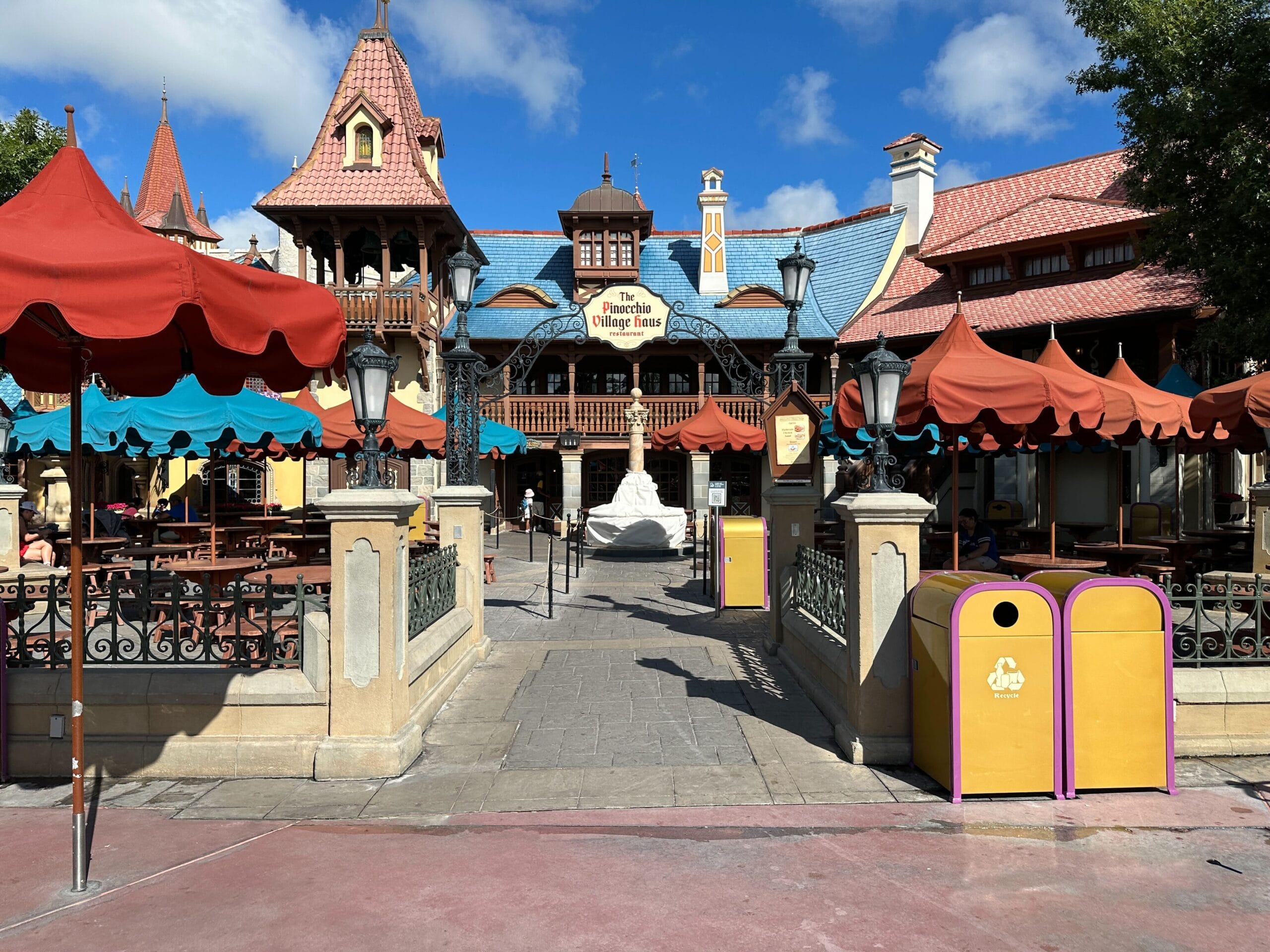 Outdoor restaurant with red and teal umbrellas near Pinocchio Village Haus, with a whimsical European-style building in the background.