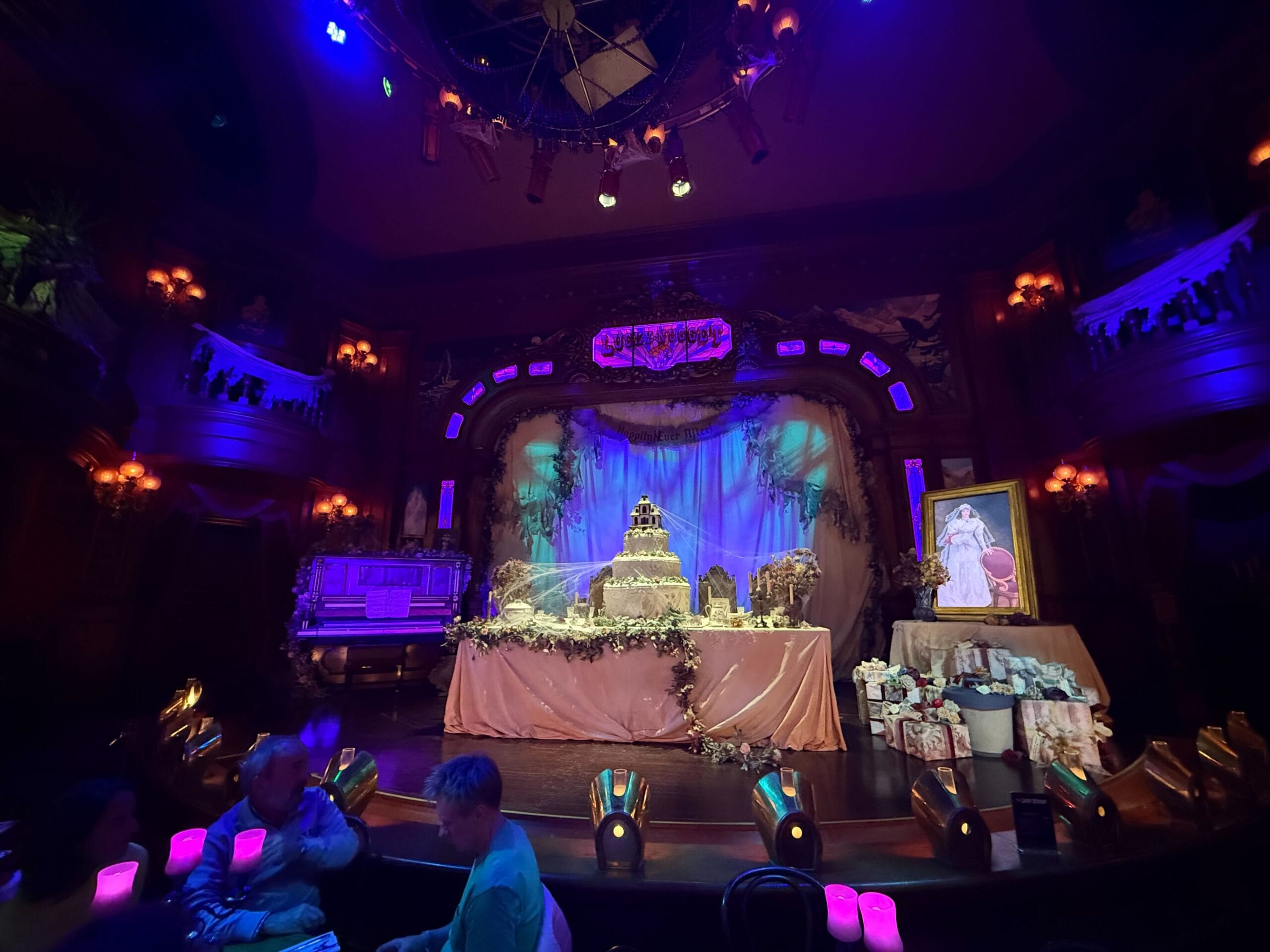 A decorated stage with a tiered cake and purple lighting evokes a celebration at Disneyland Paris’s Phantom Manor.